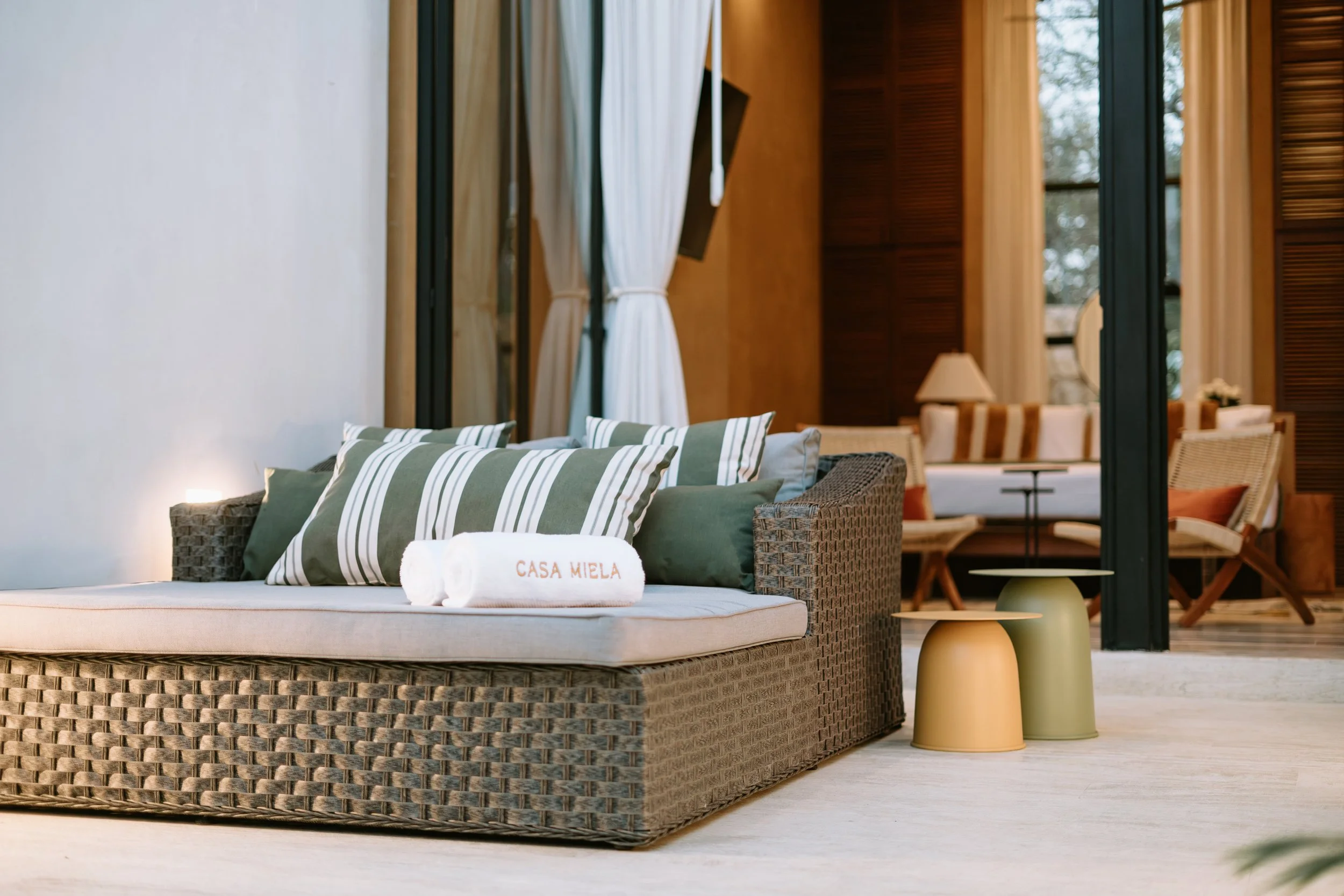 Upscale indoor hotel or resort lounge area with wicker sofa topped with striped cushions and rolled towels embroidered with 'Casa Miela'; background features a seating area with armchairs, side tables, lamps, and large windows with curtains.