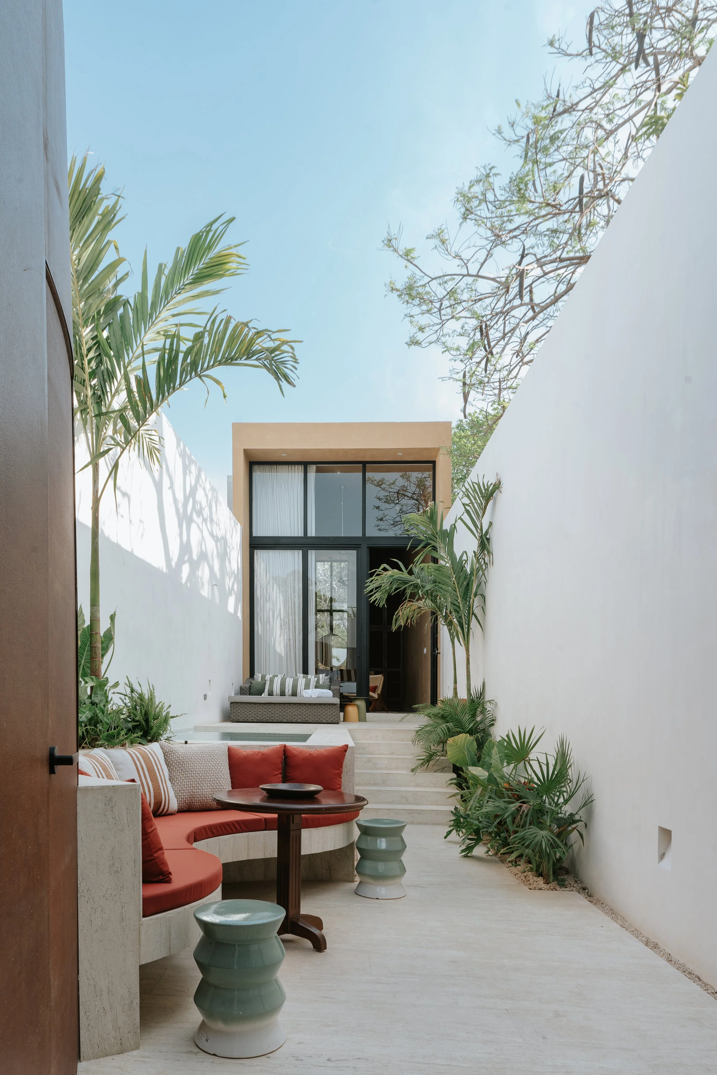 Outdoor patio with built-in seating, red cushions, potted plants, and a modern house with large glass windows in the background, under a clear blue sky. colonial house merida, restoration project