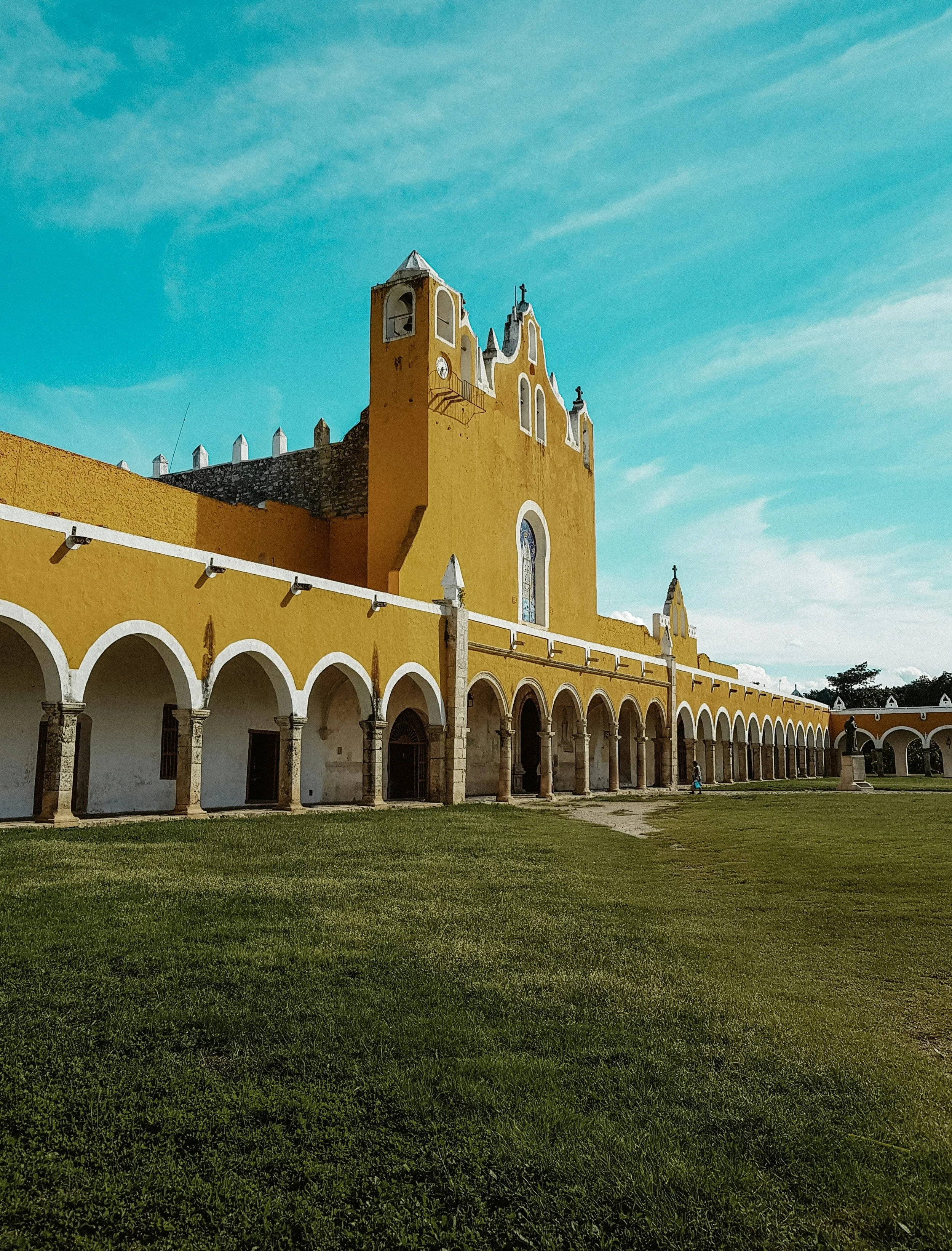 A yellow church with a tall bell tower and arched colonnade, set against a blue sky with clouds. Izamal, yucatan