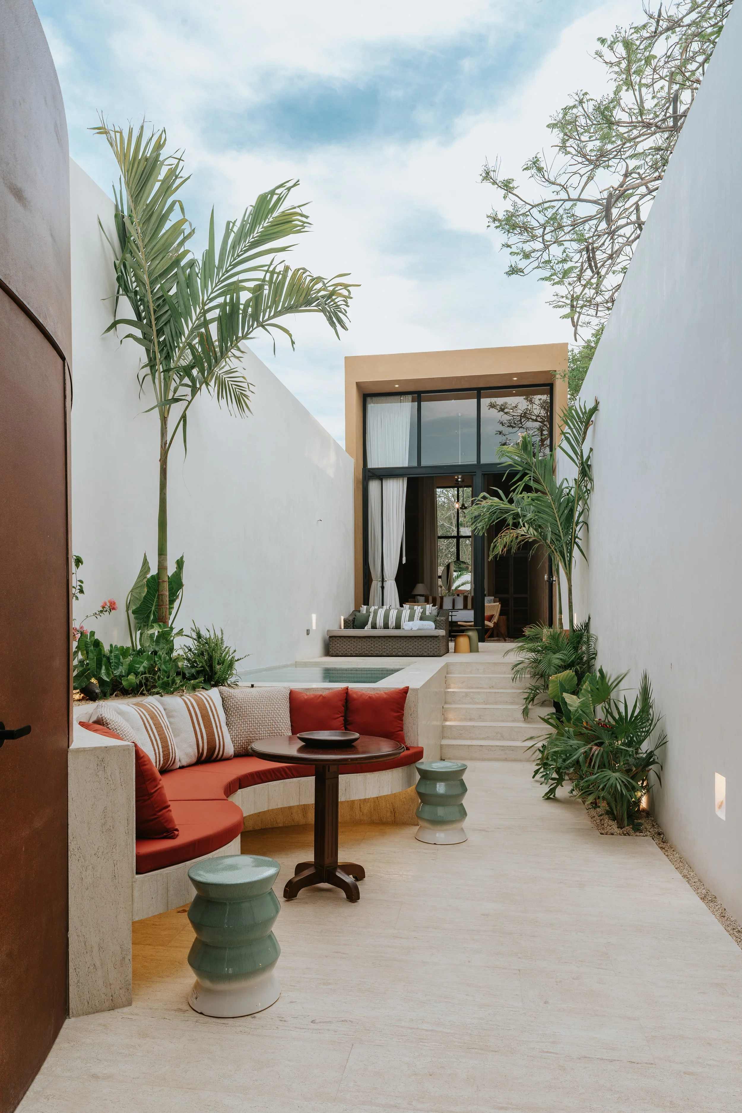 An outdoor patio area with a built-in curved bench with cushions, a round wooden table, and two ceramic stools. There are plants along the white walls, and a stepping area leads to a glass door with curtains, revealing a modern interior space.
