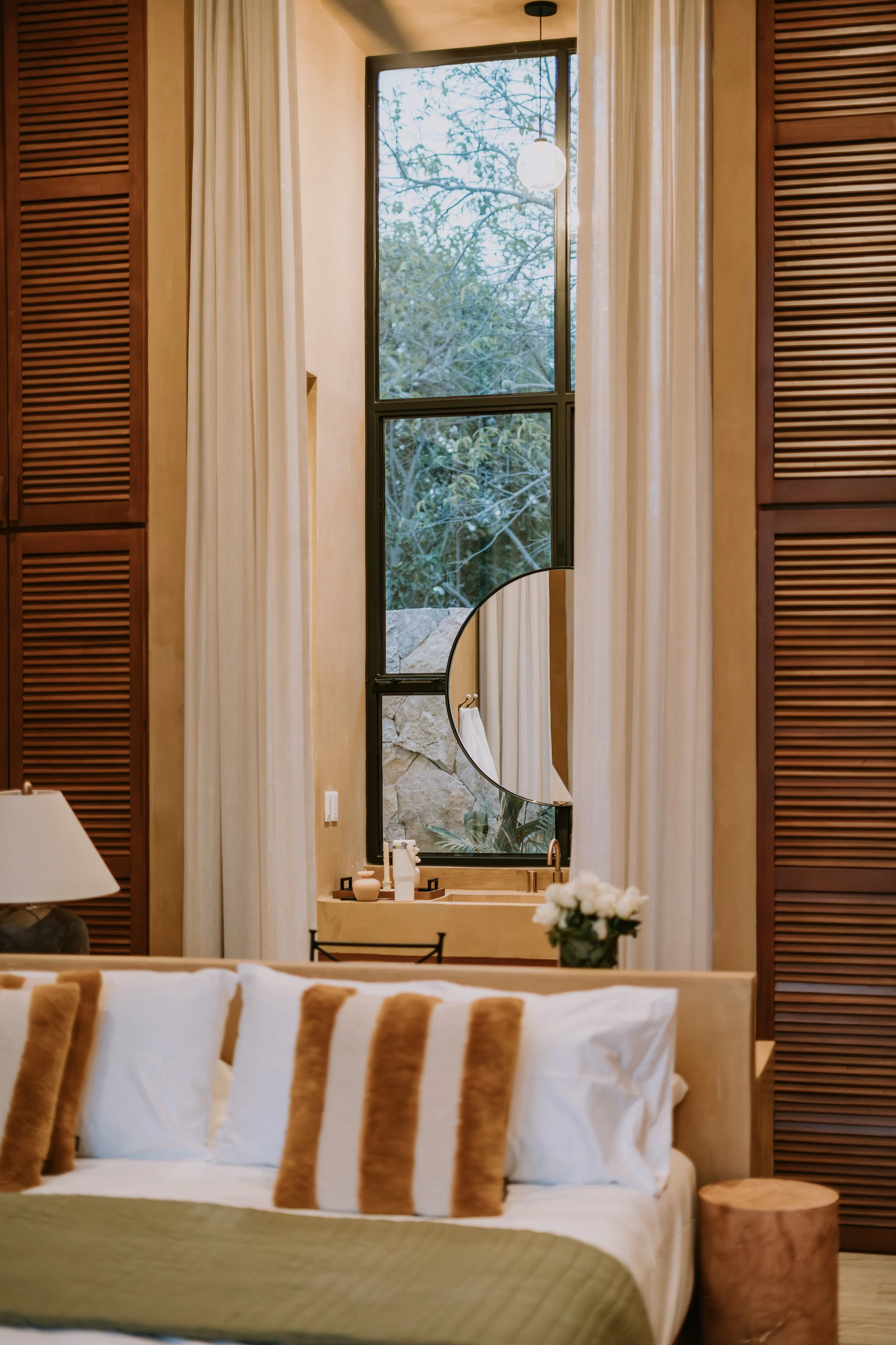 A cozy bedroom with a large window, white curtains, a round mirror, a table with decorative items, and a bed with pillows and a blanket.