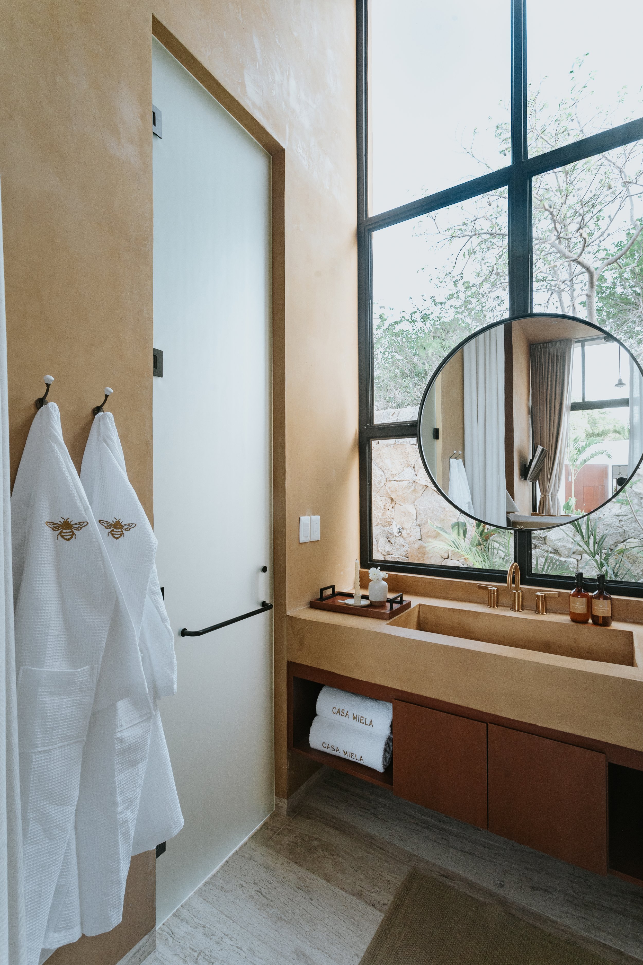 Bathroom with beige vanity, round mirror, large window with trees outside, two white towels with bee emblem, brown bottles on sink, small white vase on tray, natural light.