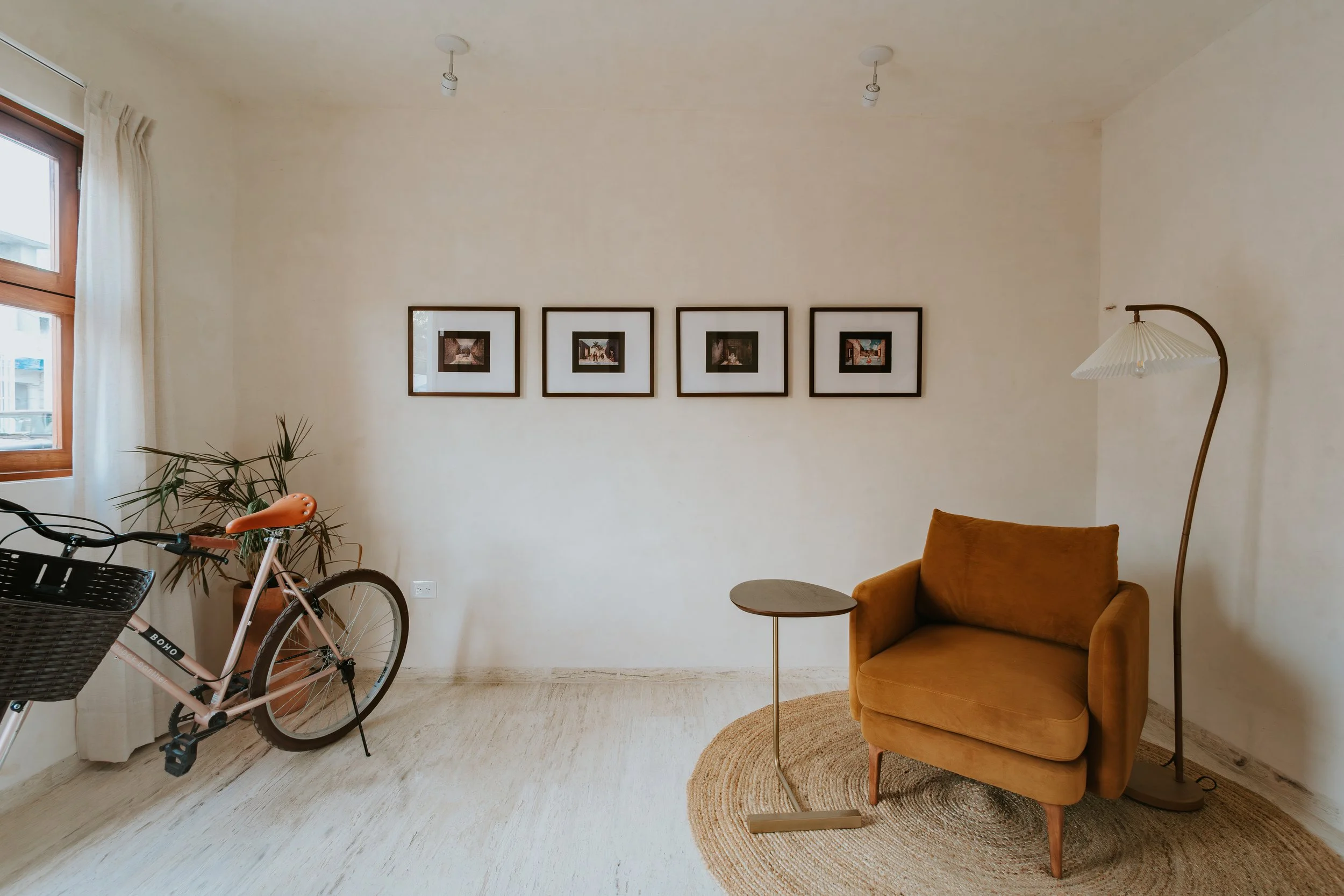Living room with a large window, a pink bicycle with a black basket, a mustard yellow armchair, a tall curved floor lamp, a small side table, a circular rug, and four framed pictures on the wall.