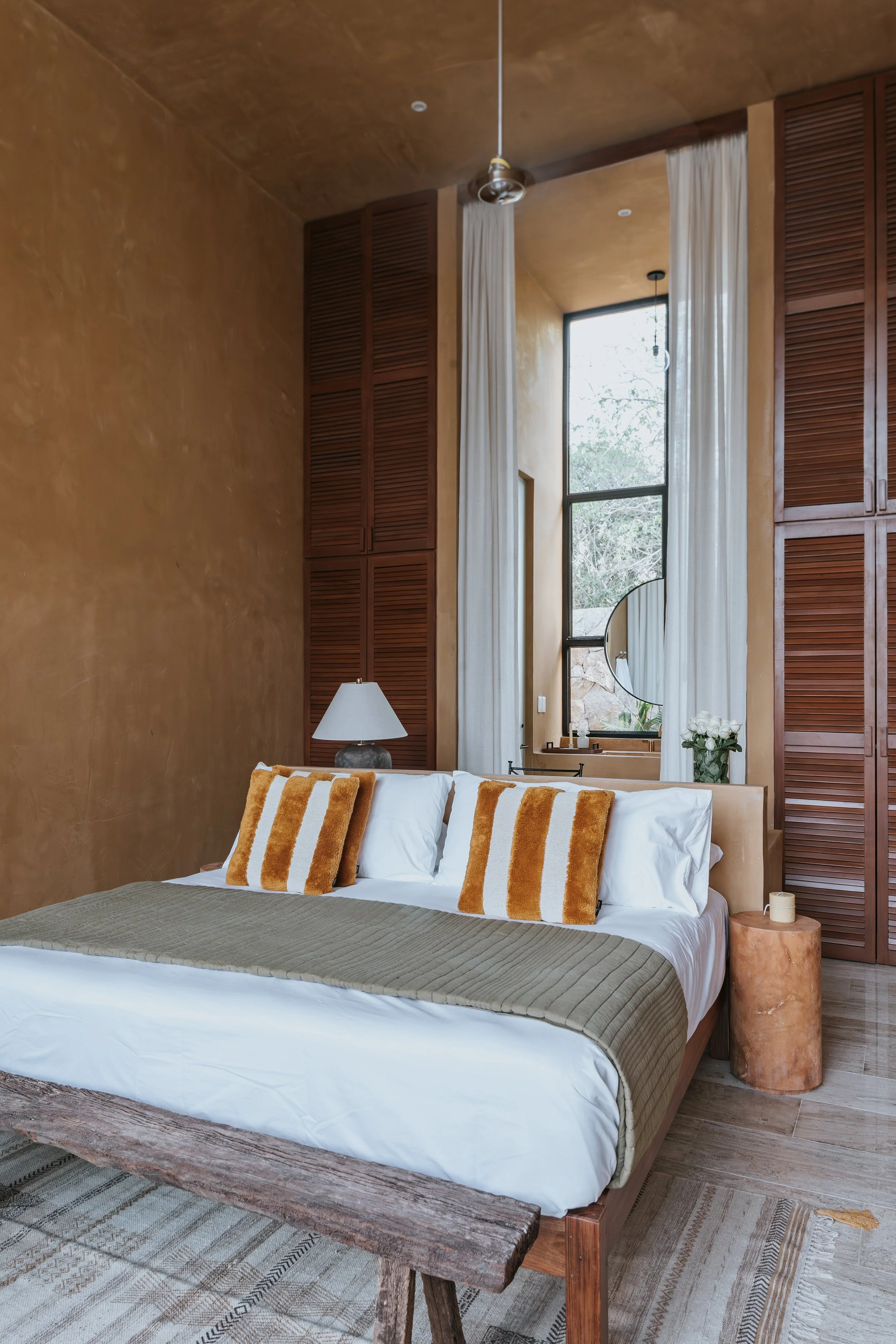A bedroom with a high ceiling, a large bed with white linens, an olive-green blanket, and striped mustard and white pillows. There is a table with a lamp on one side of the bed and a small round side table on the other. Large floor-to-ceiling curtains hang beside a tall window, and a mirror and vase with white flowers are visible in the background.