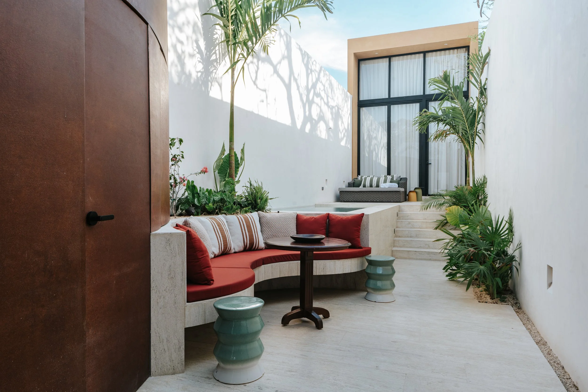 Outdoor patio with built-in curved bench with red cushions and striped pillows, small round table, and two green ceramic stools, surrounded by white walls, green plants, and a modern building with large glass doors in the background. colonial house m