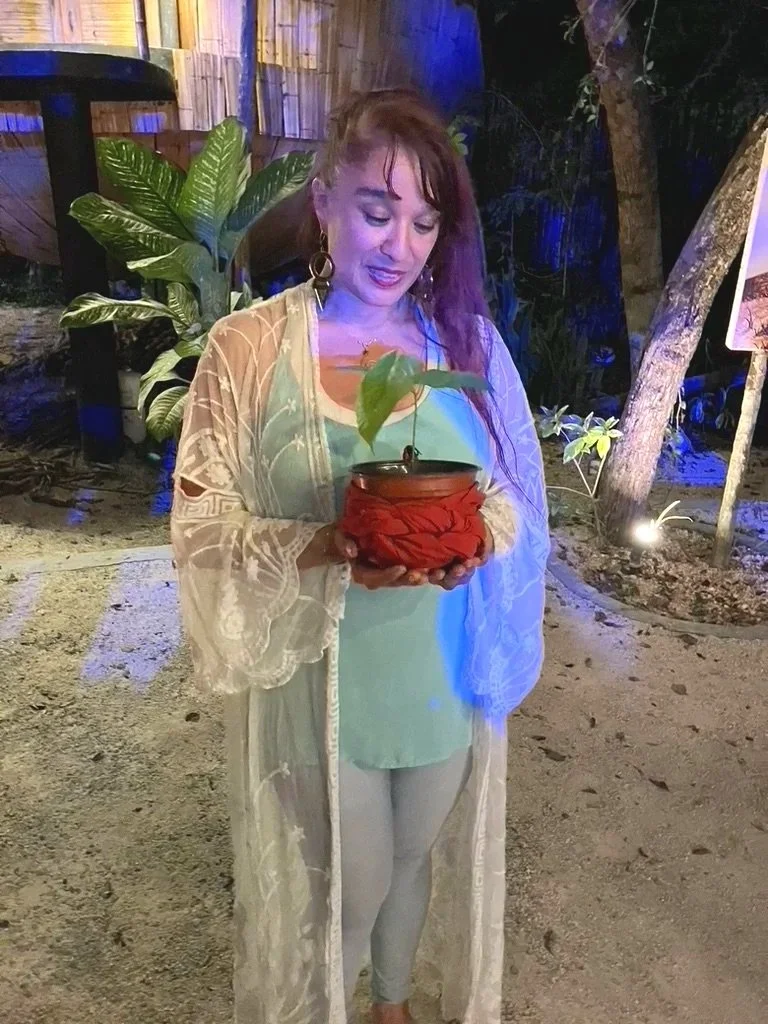 A woman holding a small potted plant with a large leaf, standing outdoors at night with plants and trees in the background, illuminated by blue and purple lighting.