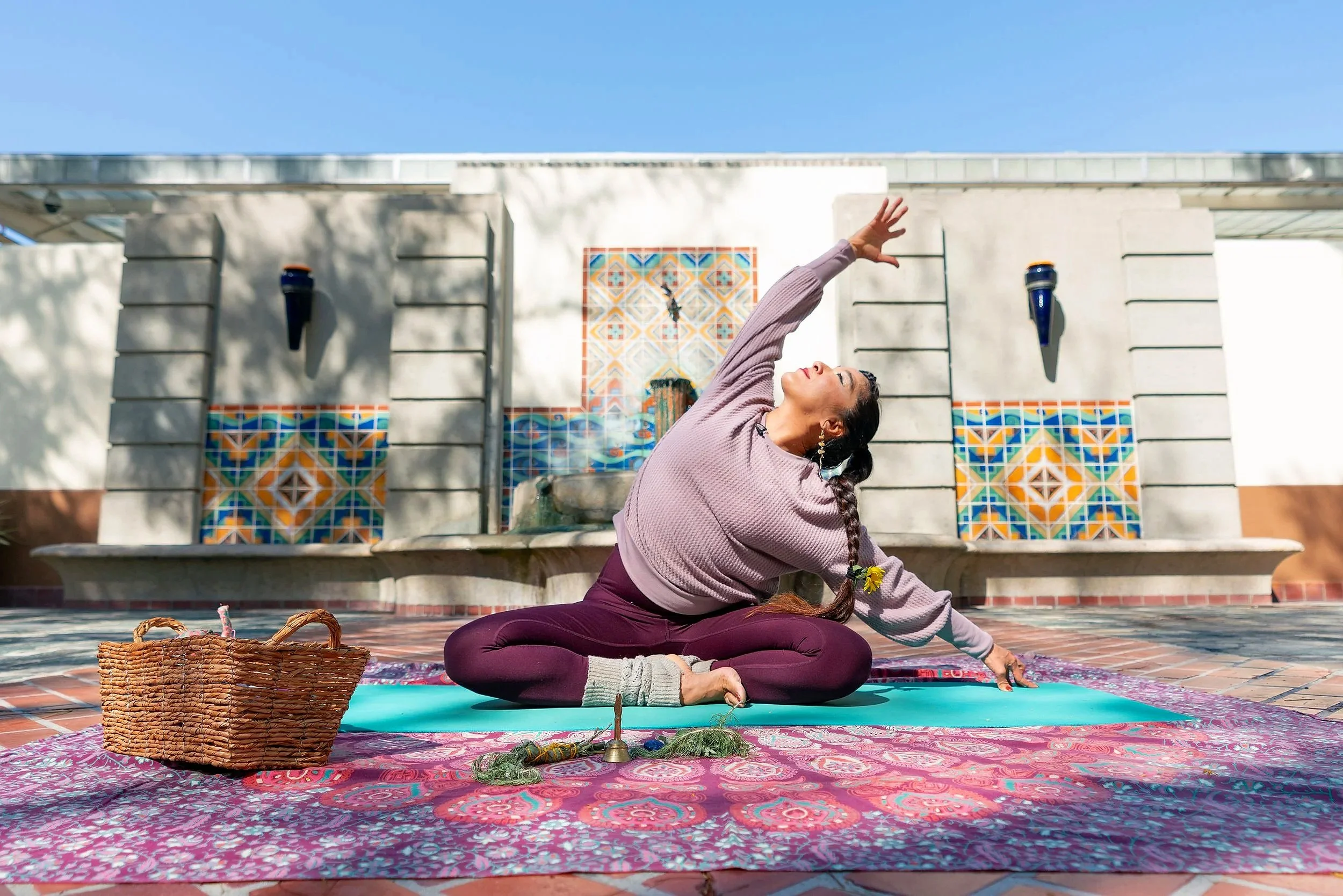 A woman practicing yoga outdoors on a blue mat, sitting cross-legged on a pink and purple patterned rug, with her left arm extended upward and her head tilted back. She has a braid with a yellow flower. There is a wicker basket and small bells with greenery in front of her, and a decorative water fountain behind her, with colorful tile accents and a wall-mounted water feature. Clear blue sky is overhead.