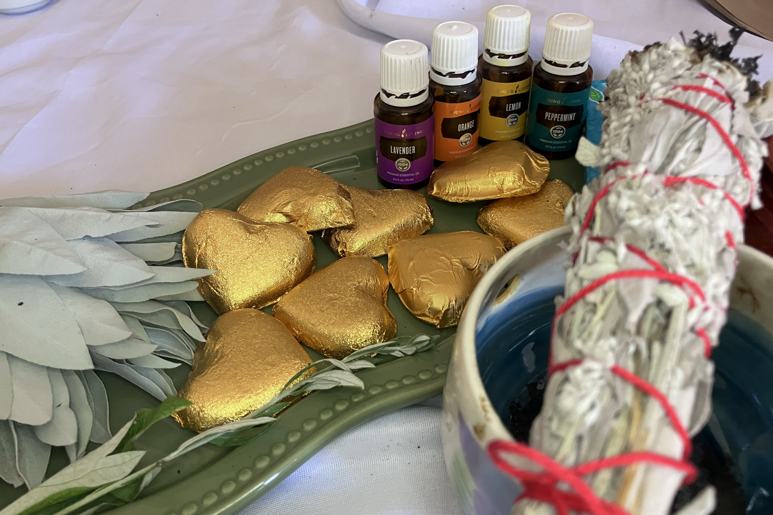 A table with a green tray containing gold-wrapped heart-shaped chocolates, herbal leaves, and small bottles of essential oils in lavender, orange, lemon, and peppermint. Part of a lava lamp with red rubber bands around the top is visible.