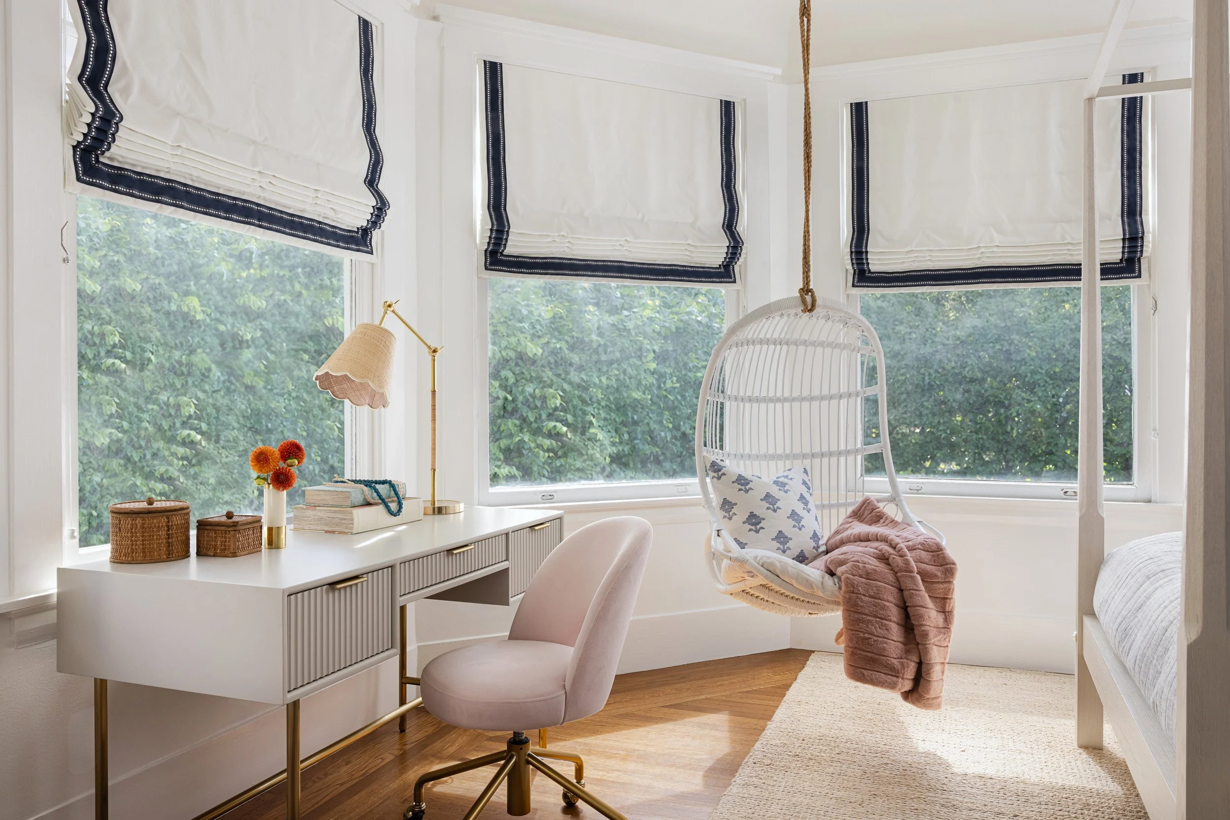 home office with white desk, hanging rattan chair and navy-trimmed roman shades