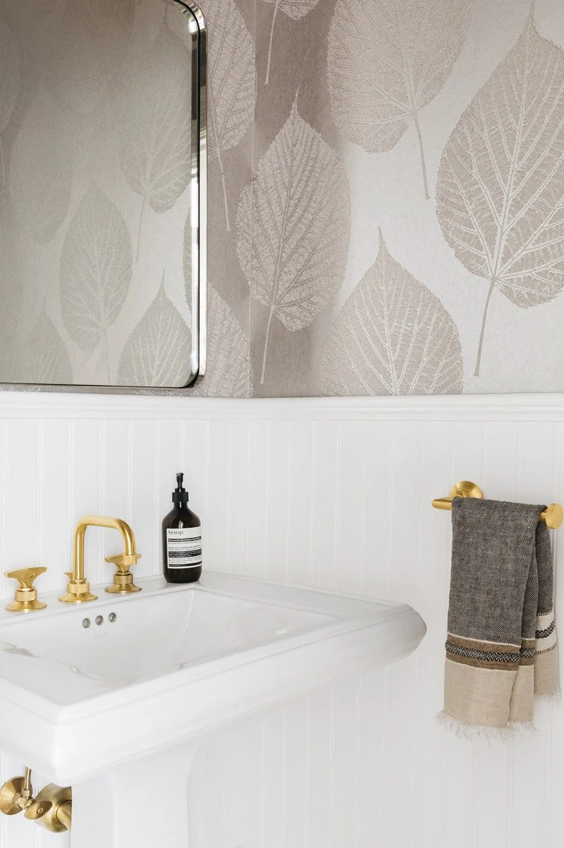 pedestal sink with brass fixtures beneath leaf print wallpaper