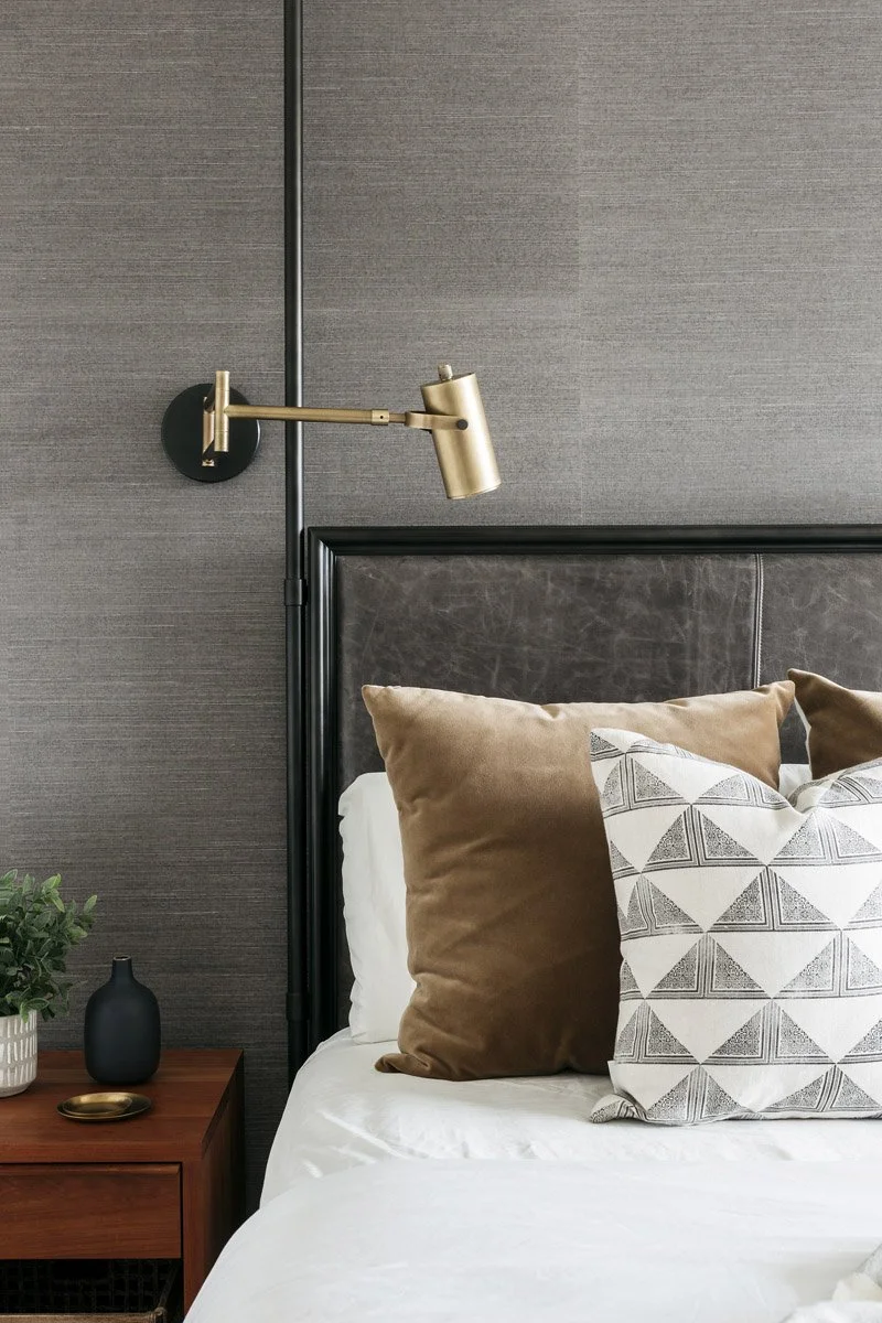 bedside detail with brass swing-arm sconce, leather headboard and patterned pillows