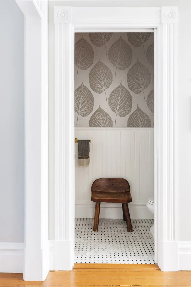 view into a wallpapered powder room with a wooden stool