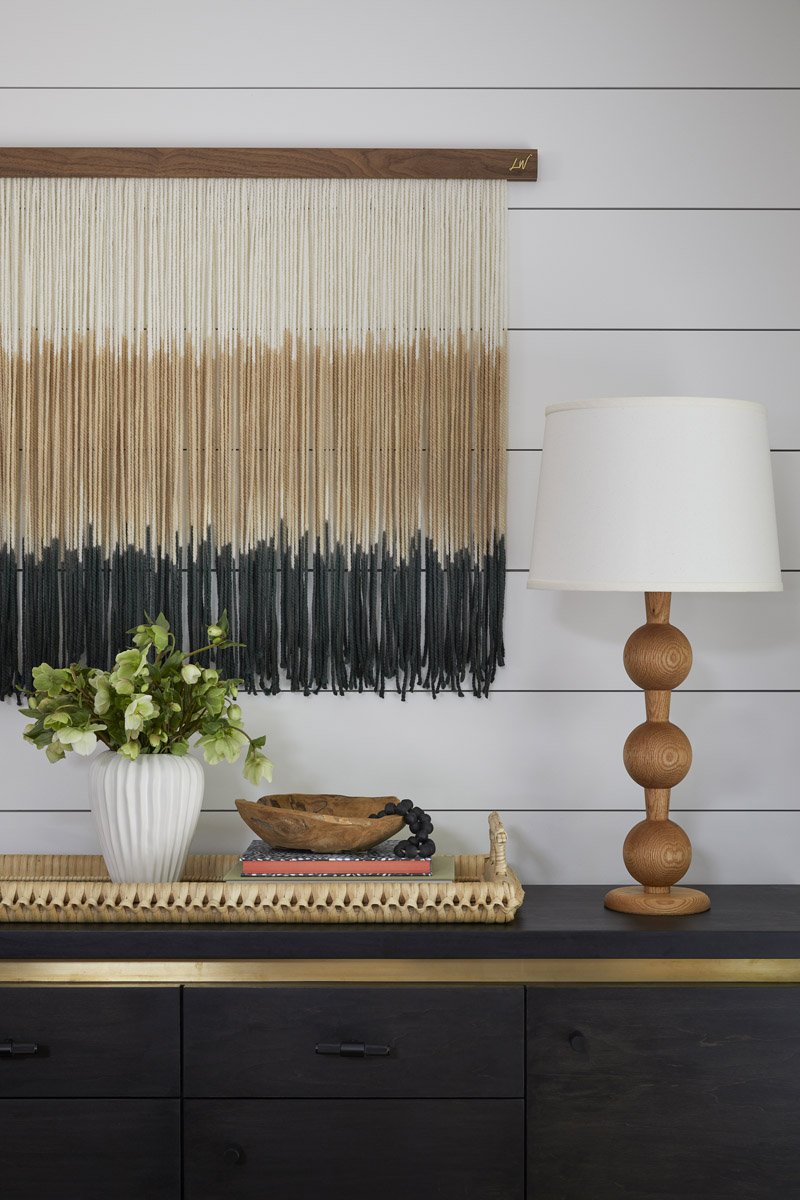woven wall hanging above a black sideboard with a turned wood lamp
