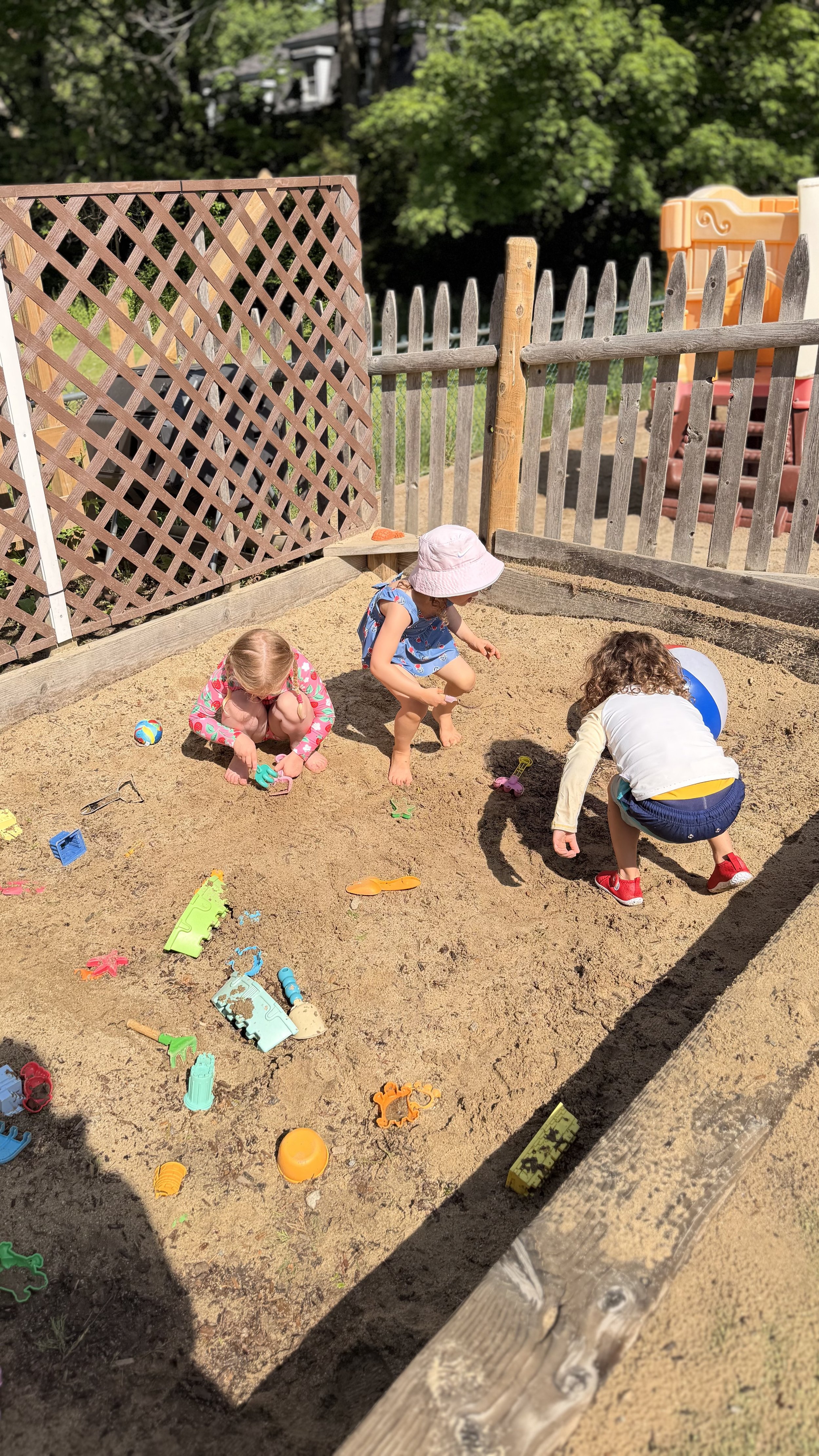 Three young children play together in a sandbox filled with toys on a sunny day at an outdoor playground.