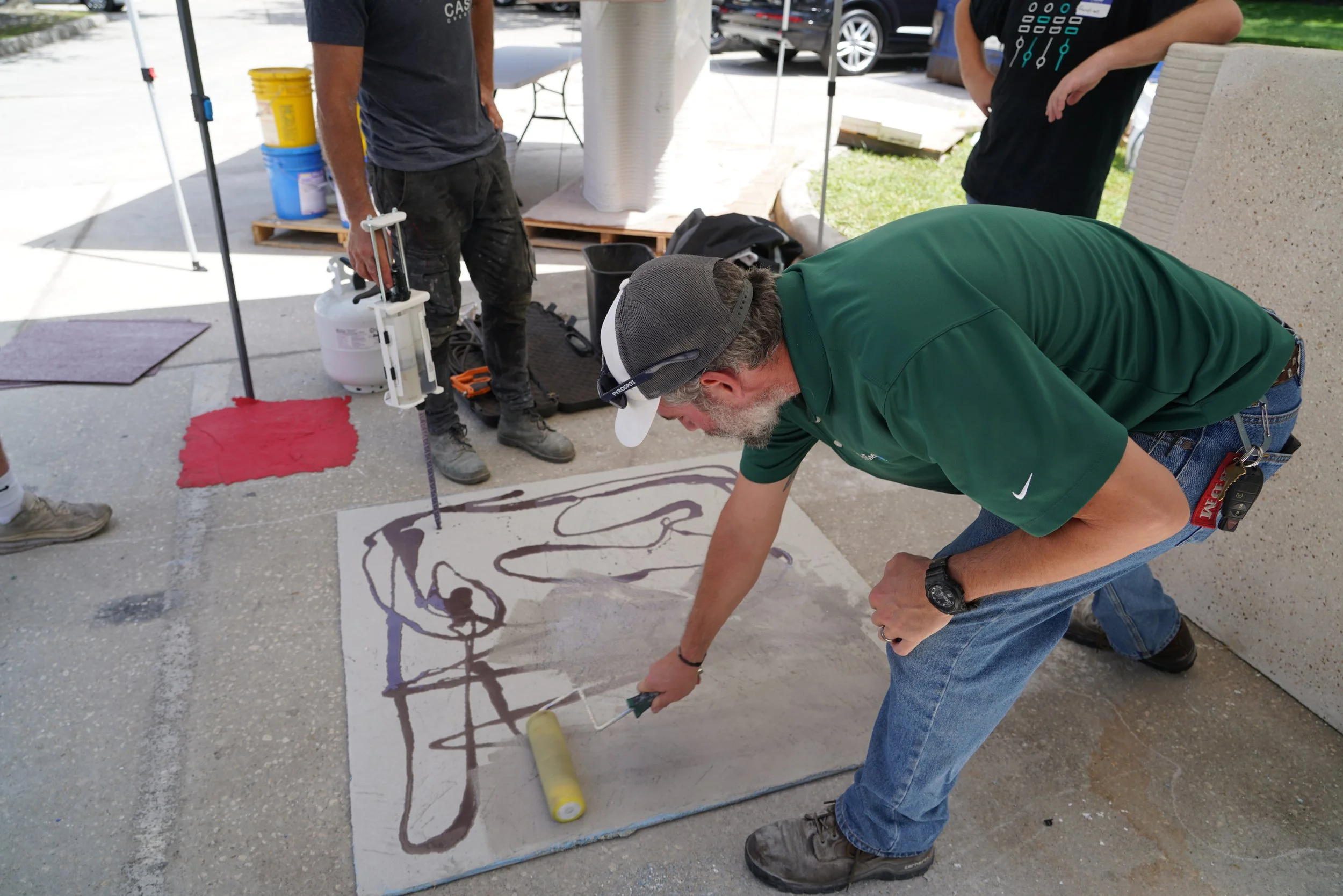 A man is painting a large abstract line art on a white surface outdoors, using a roller brush. Other people stand nearby, with one man holding painting supplies, and a tent and parked cars are in the background.