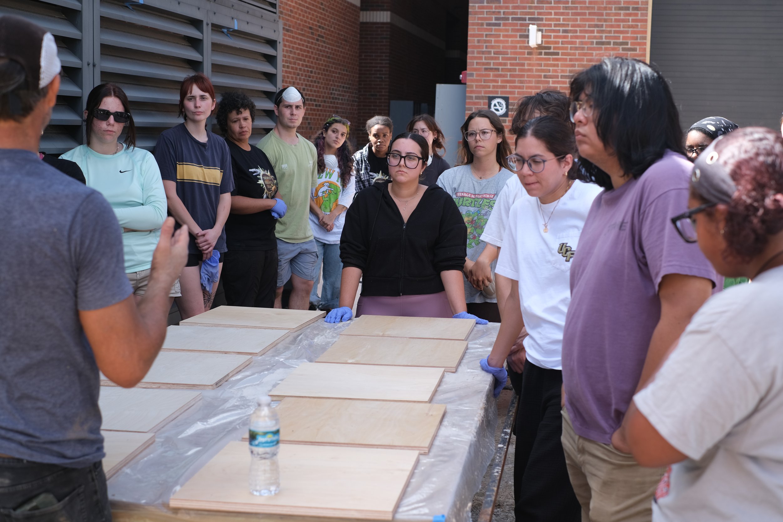 A group of people gathered around a table with wooden boards, listening to a person who is speaking, at an outdoor event.