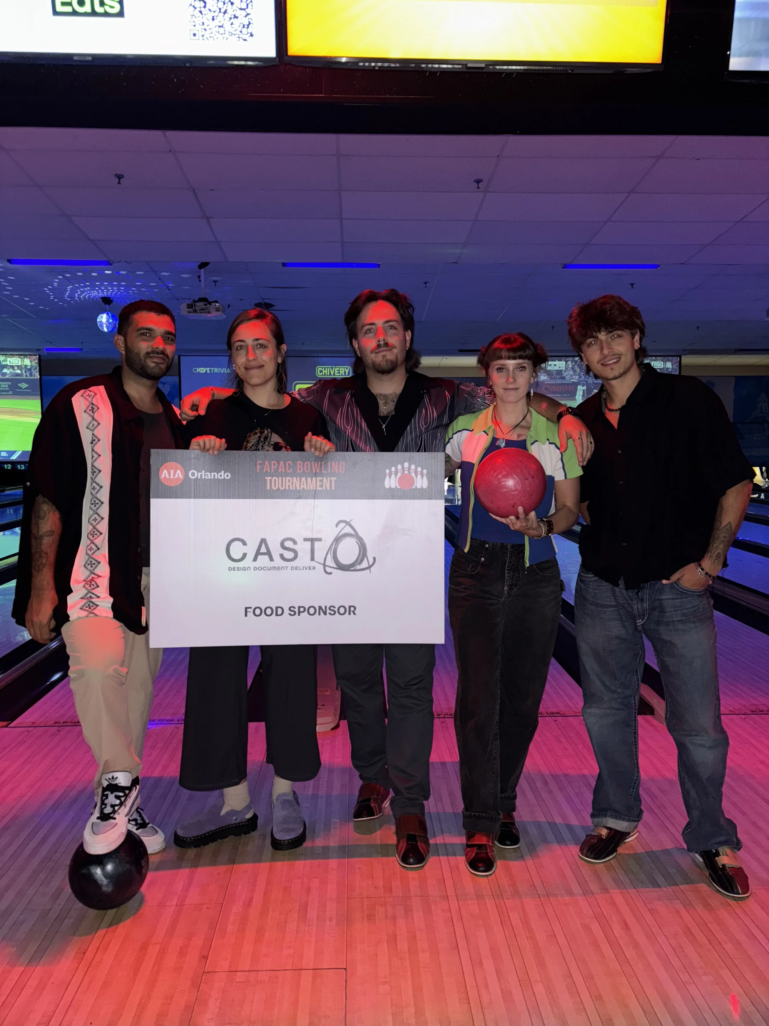 Group of five people at a bowling alley holding a large sponsorship check, with one person holding a red bowling ball.