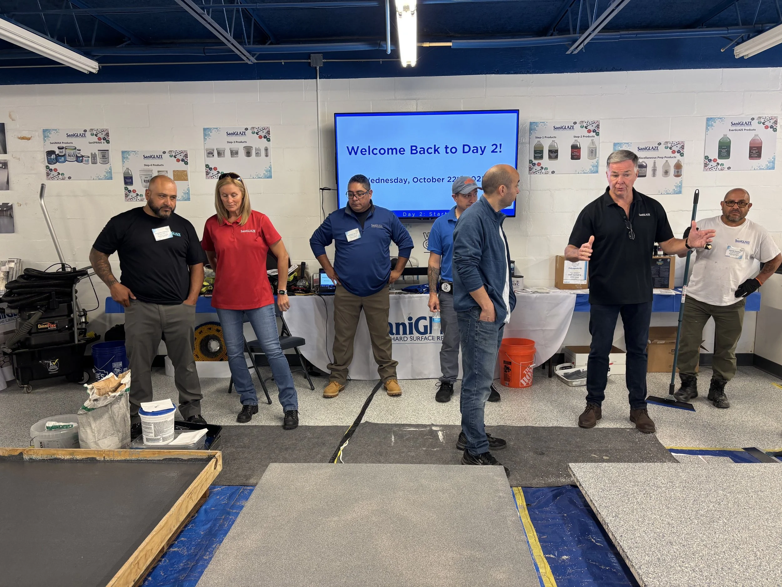 Group of seven people standing in a room in front of a large screen that reads 'Welcome Back to Day 2! Wednesday, October 22, 2022'. They are engaged in conversation, with some looking at each other and others looking at the camera. The room appears to be a workshop or conference setting, with various tools and equipment on tables and walls.