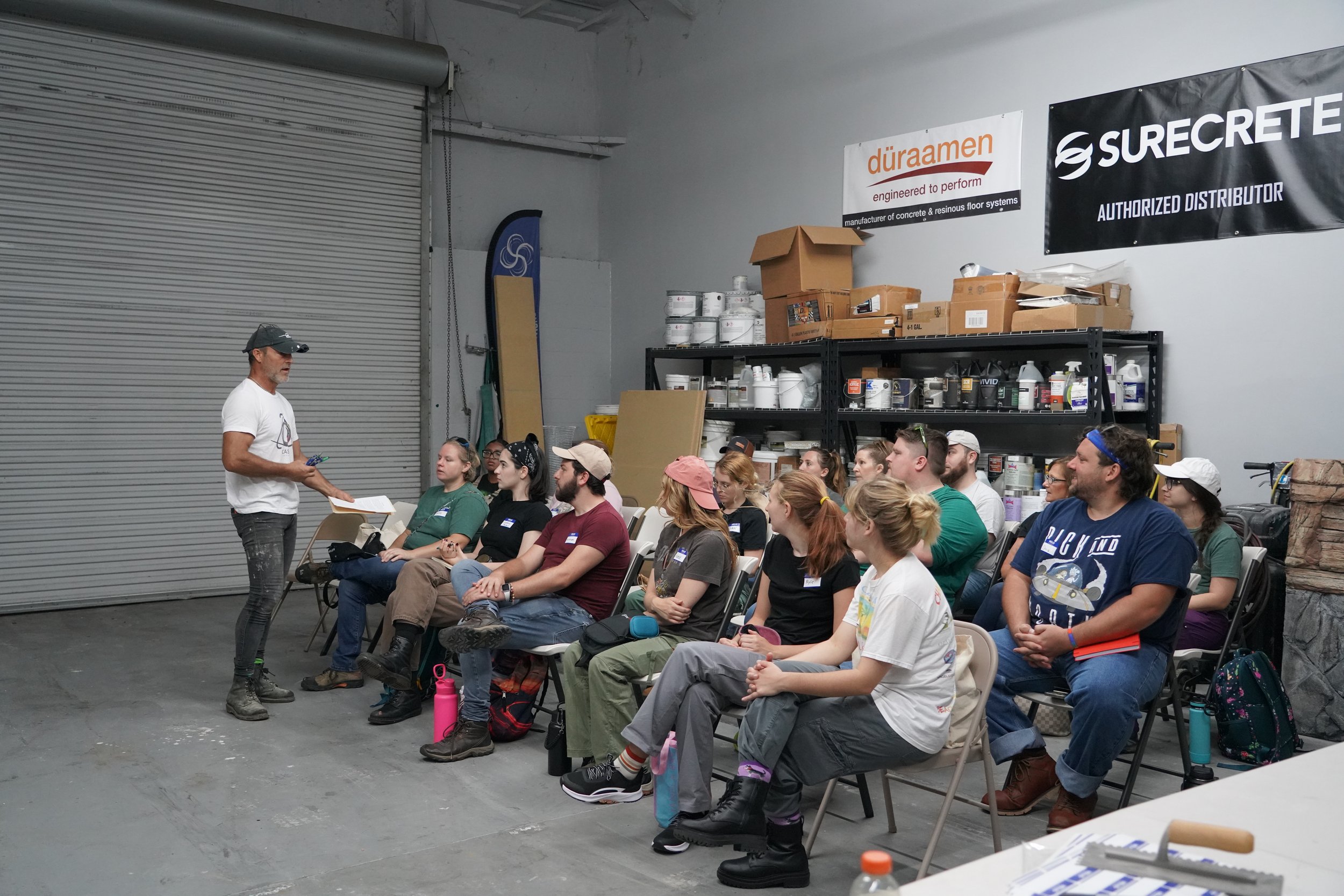 A man is giving a presentation to a group of people seated in a warehouse or workshop setting, with shelves of supplies and banners on the wall behind them.