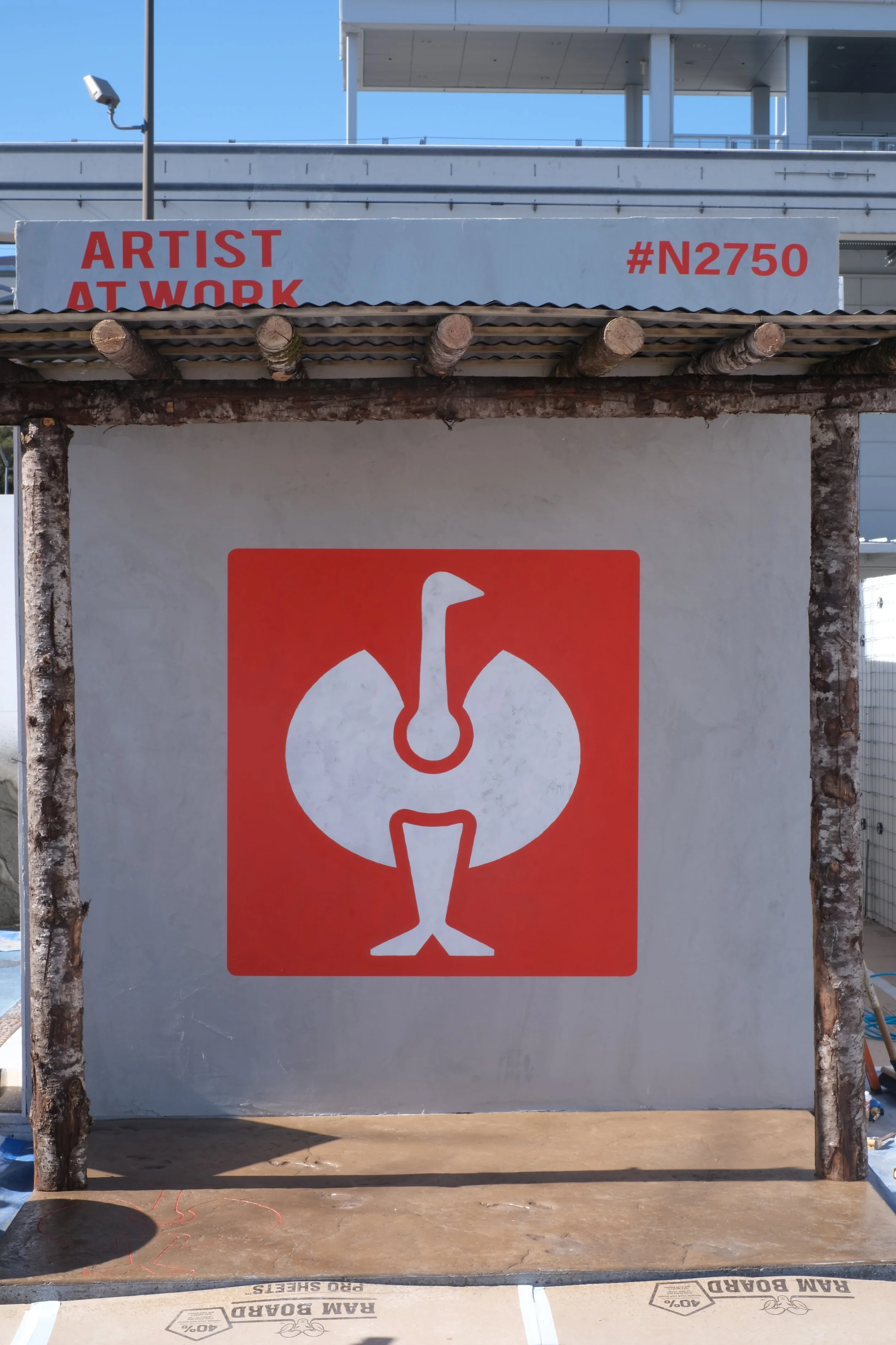 A construction site with a sign that reads 'ARTIST AT WORK' and a red symbol of a broken duck or bird. The scene includes a wooden and concrete structure with a blue sky background.
