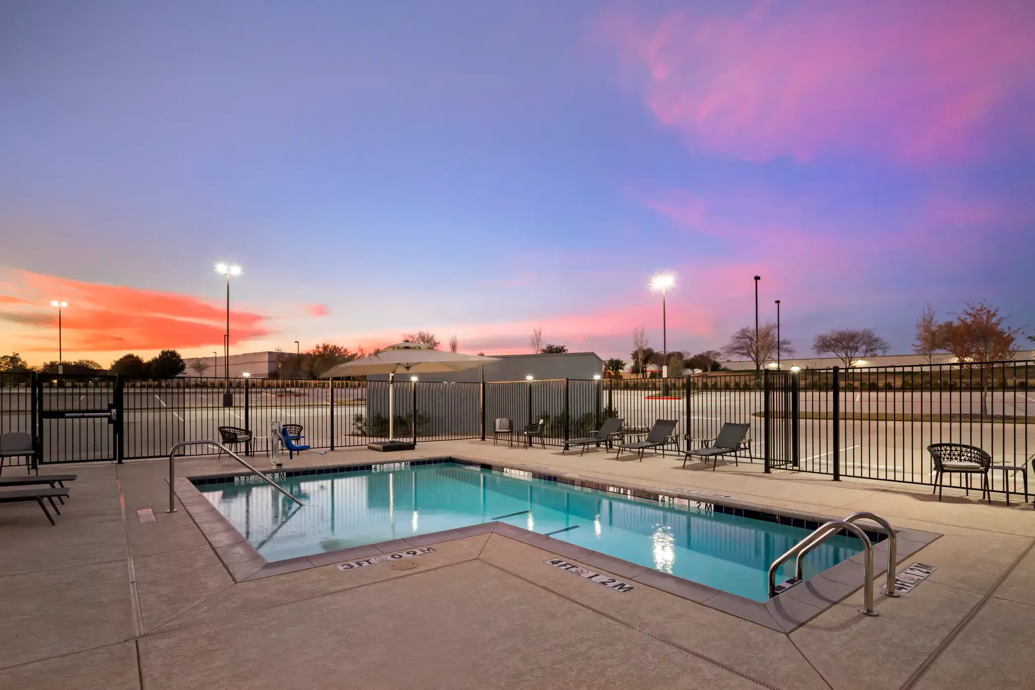 An empty outdoor swimming pool surrounded by lounge chairs and a fence at dusk, with the colorful sky and a few trees in the background.