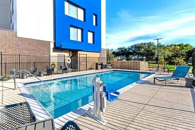 Outdoor swimming pool area with lounge chairs, a blue lounge chair, small tables, and a black fence. Modern building with blue and white exterior and windows, against a bright blue sky with some clouds.