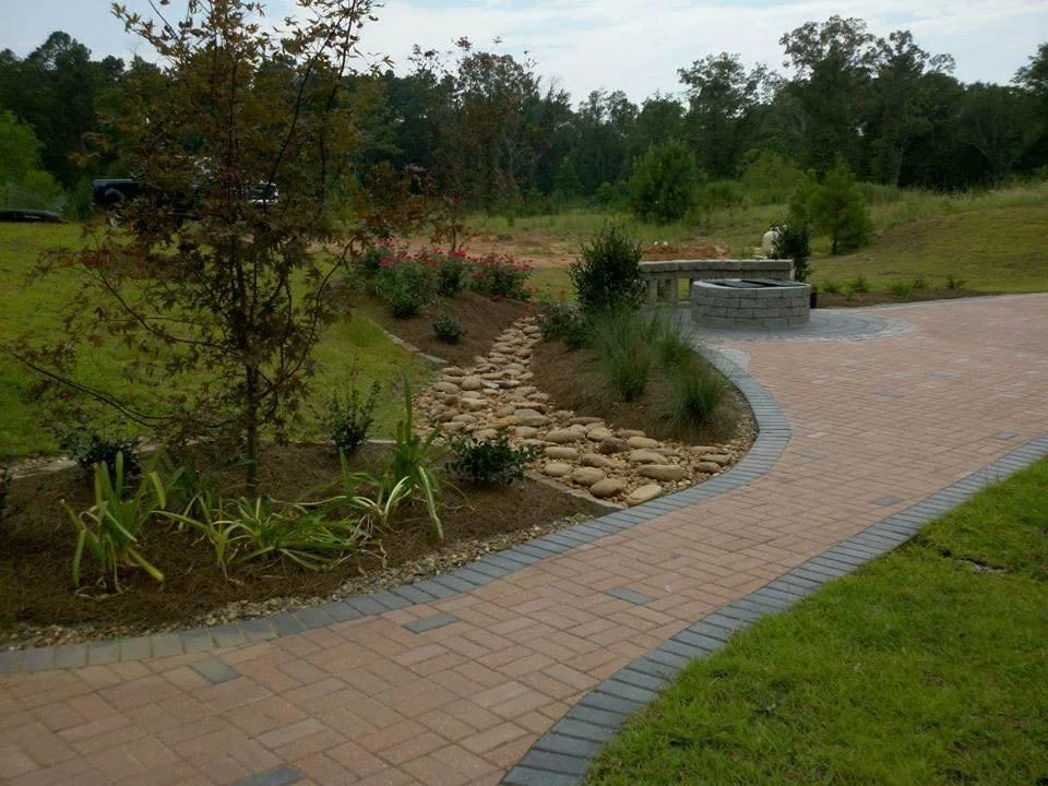 A landscaped backyard with a brick pathway, a small tree, flower bed with plants, a stone fire pit, and a grassy area with trees in the background.