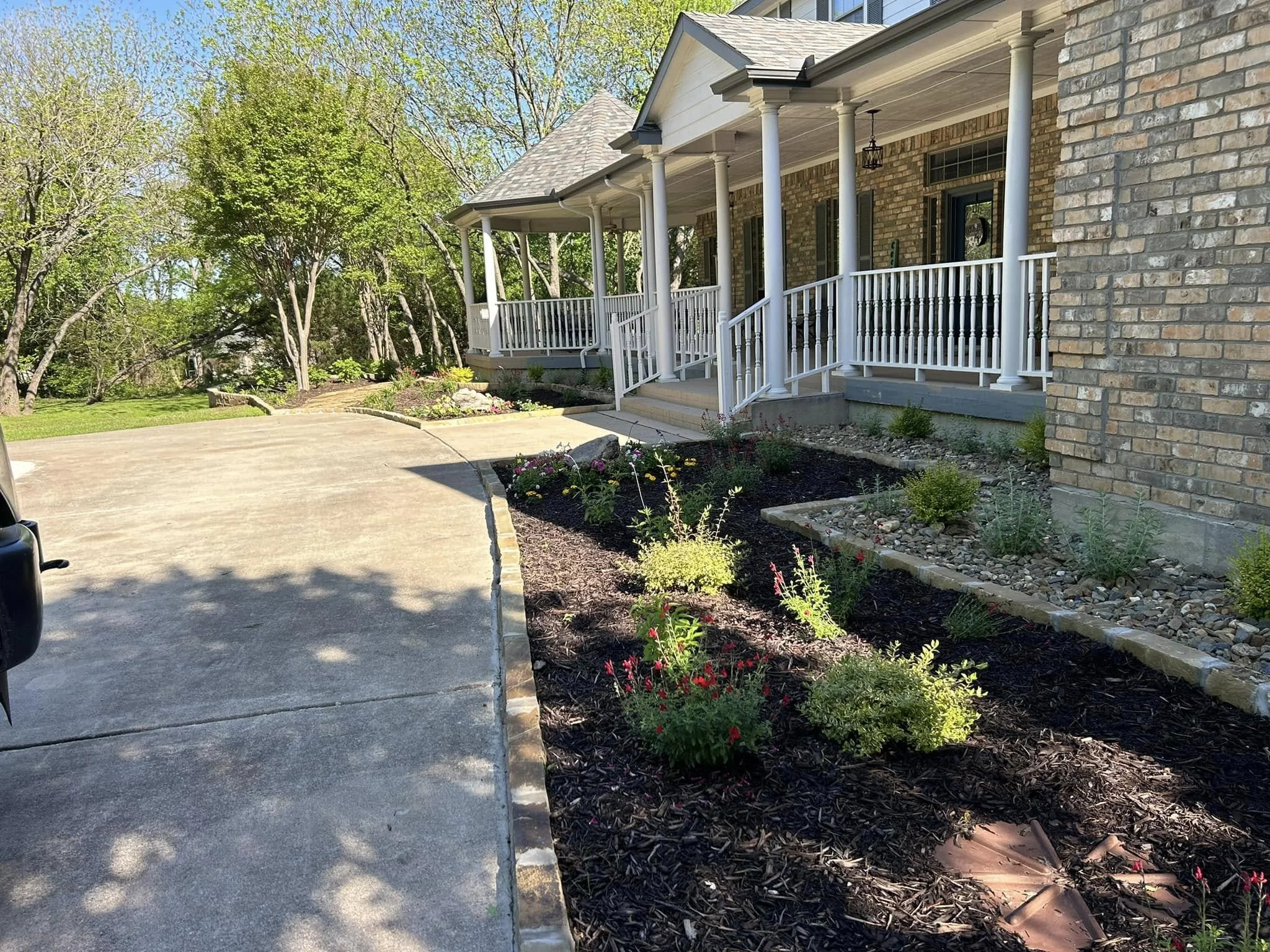 Front yard with mulch flower bed, stone pathway, and brick house with porch, columns, and white railing, surrounded by green trees and a concrete driveway.