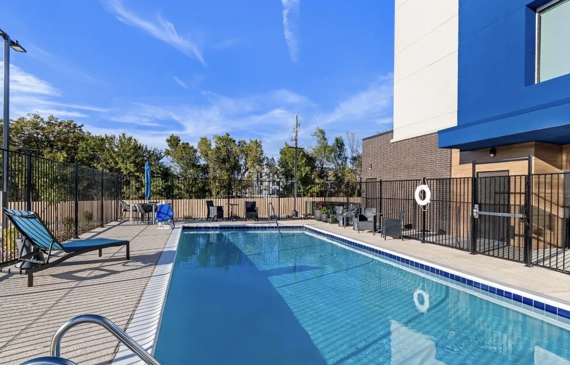 Empty outdoor swimming pool with lounge chairs, tables, and fencing on a sunny day with blue sky and trees in the background.