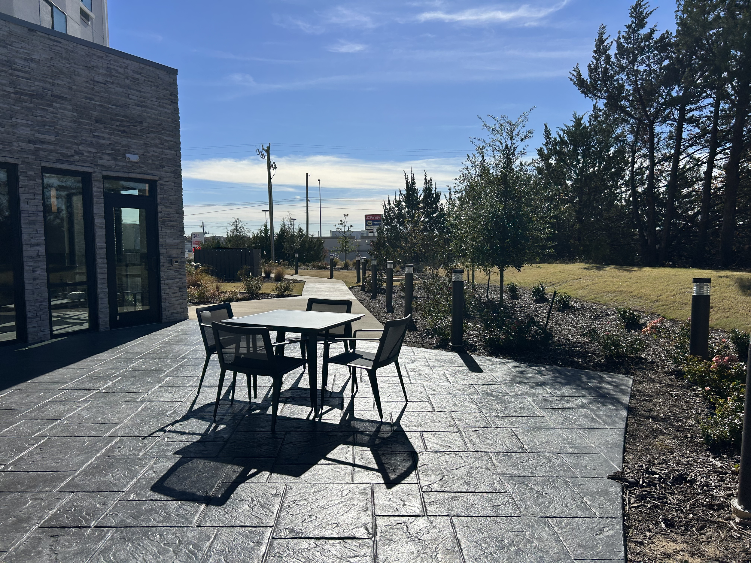 Outdoor patio with black metal table and chairs, adjacent to a building with stone exterior, surrounded by small trees and shrubs, under a blue sky with some clouds.
