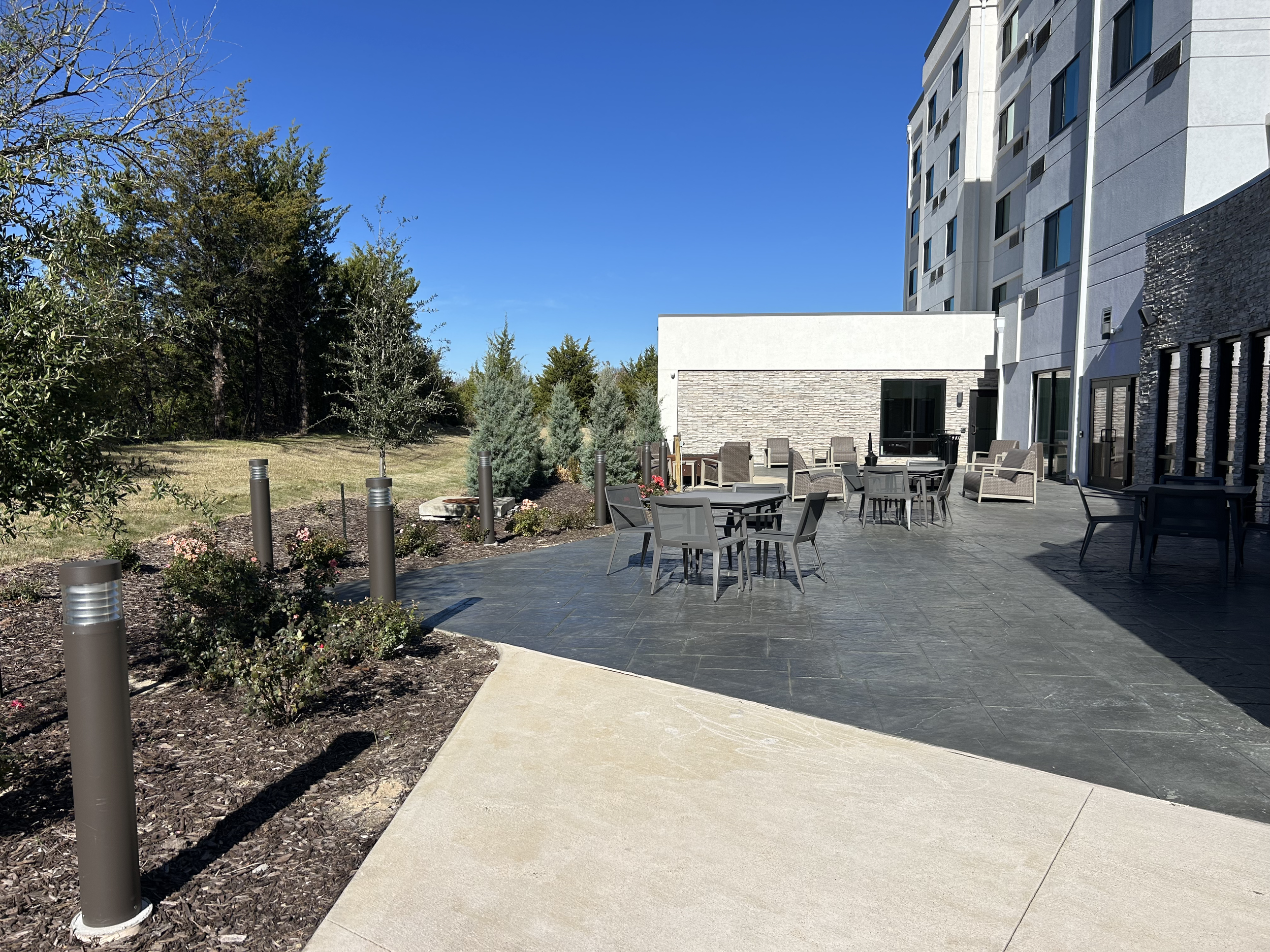 Outdoor patio with tables and chairs, adjacent to a modern multi-story building, with landscaped garden and trees in the background under a clear blue sky.