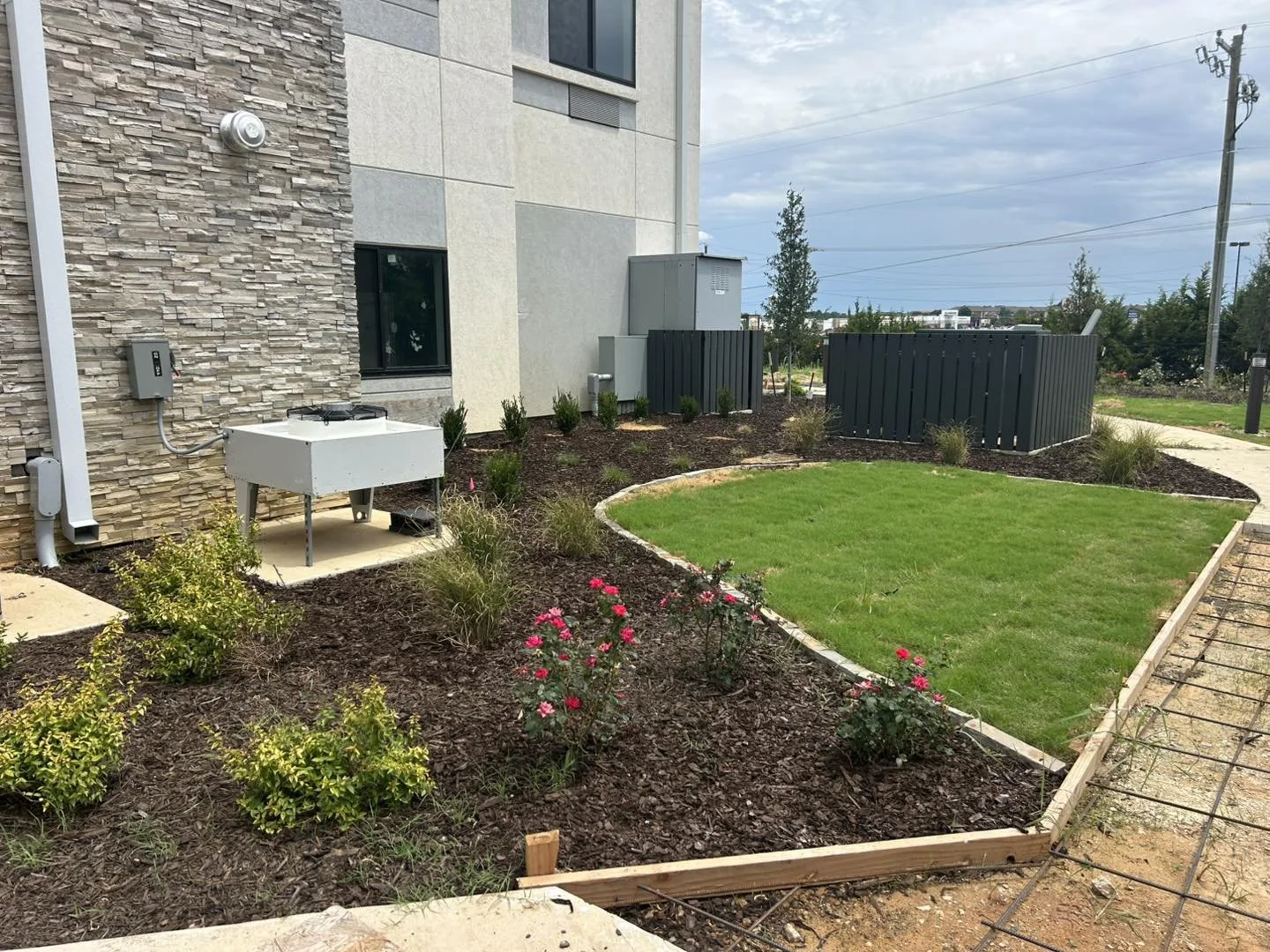 A landscaped backyard with a small grassy lawn, flower beds, and newly planted bushes and flowers, adjacent to a modern building with stone and beige siding, and utility boxes and fencing in the background.