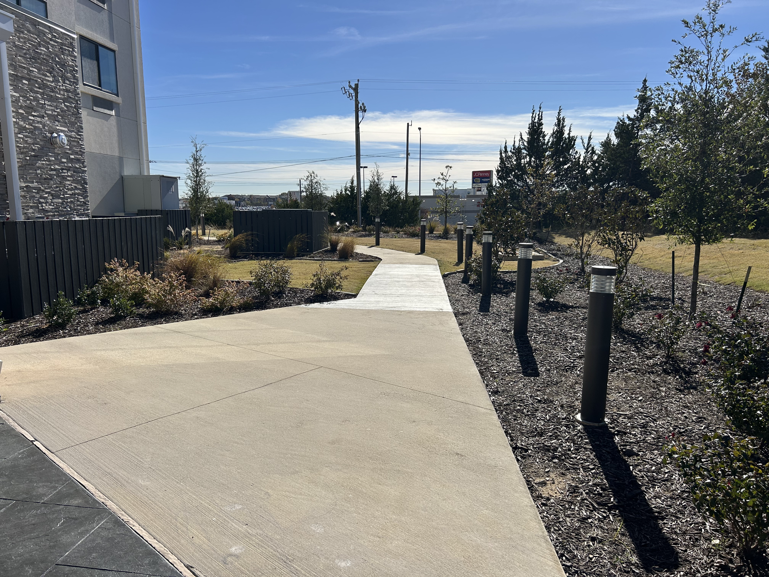 A sidewalk in a landscaped area with small bushes, trees, and evenly spaced black bollard lights, next to a modern building with a stone and concrete exterior, under a blue sky with some clouds.