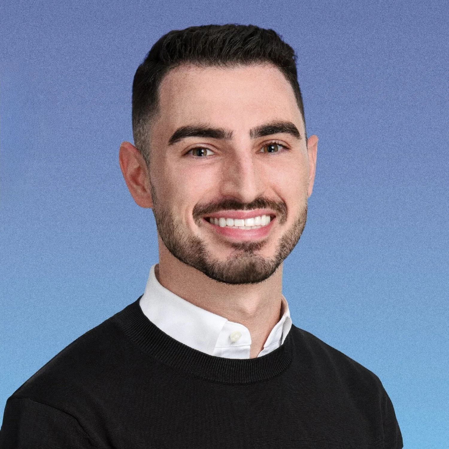 A headshot of a young man with dark hair, beard, and mustache, smiling, wearing a white shirt and a black sweater, against a blue background.