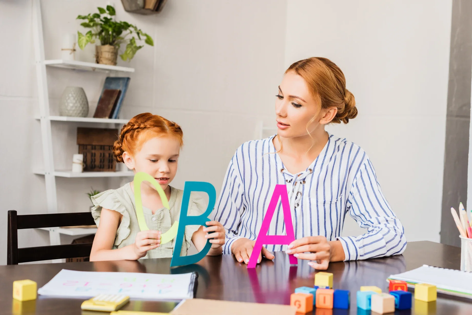 A woman and a girl sitting at a table with colorful alphabet letters, blocks, and notebooks, engaging in educational activities.