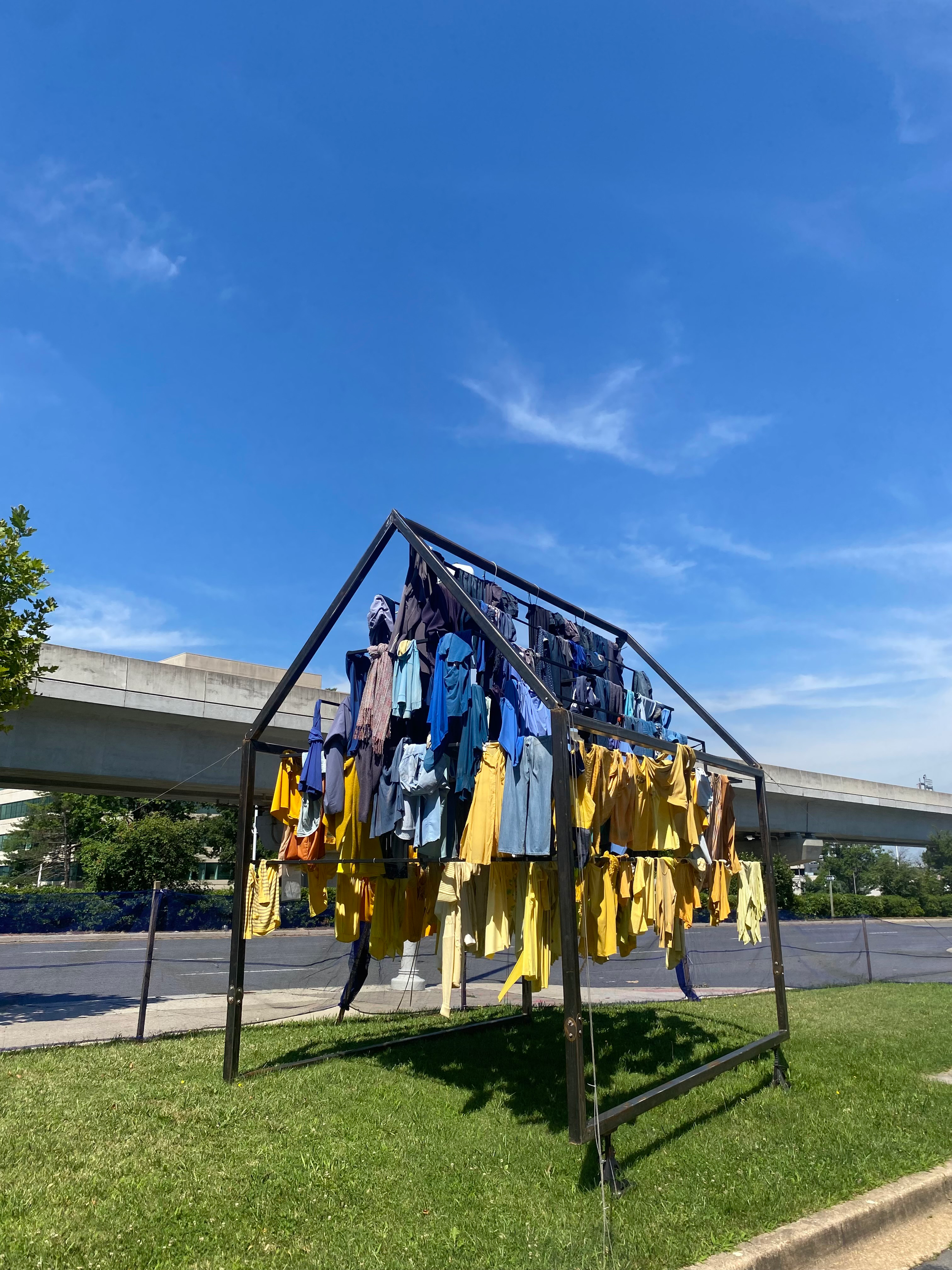 Steel house frame on grass with clothes hanging off it in shades of yellows and blues