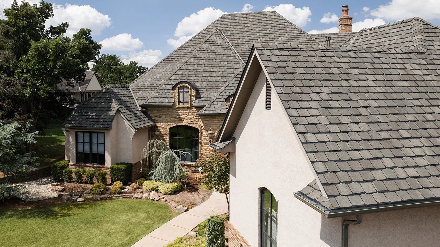 A two-story house with a stone and stucco exterior, dark gray shingle roof, and a landscaped front yard with shrubs, trees, and a walkway.