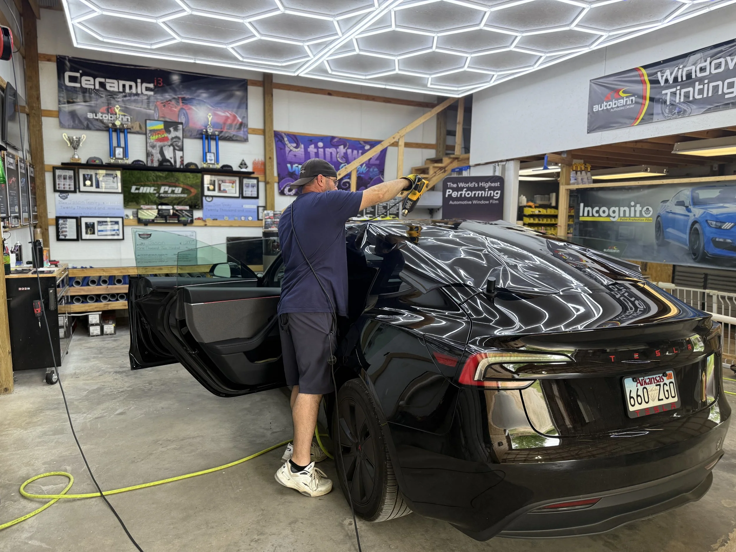 A man working on the roof of a black Tesla in a garage workshop, with car doors open and various banners and awards on the walls.