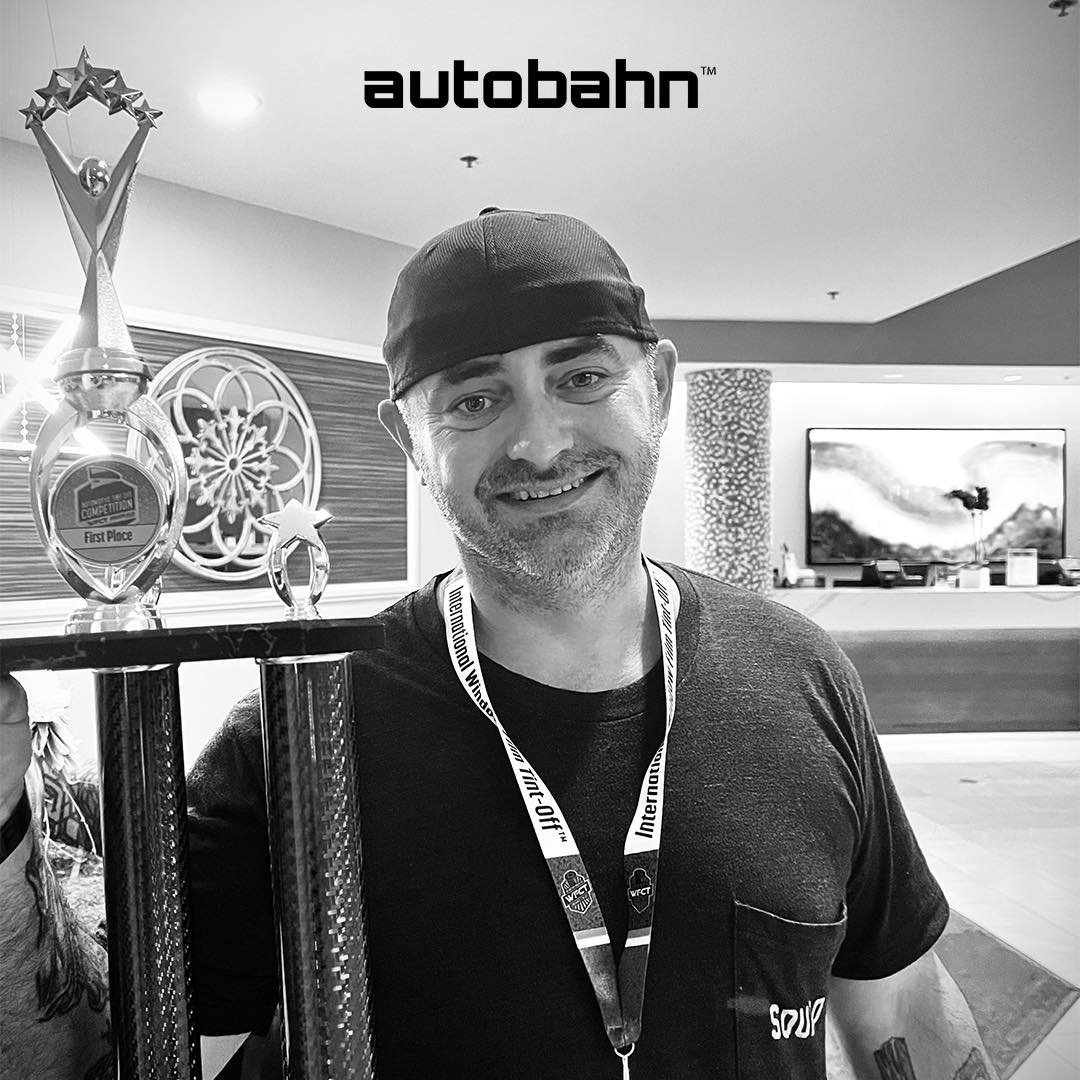 Person smiling holding a large first place trophy indoors, wearing a cap and a lanyard, with "autobahn" logo above.