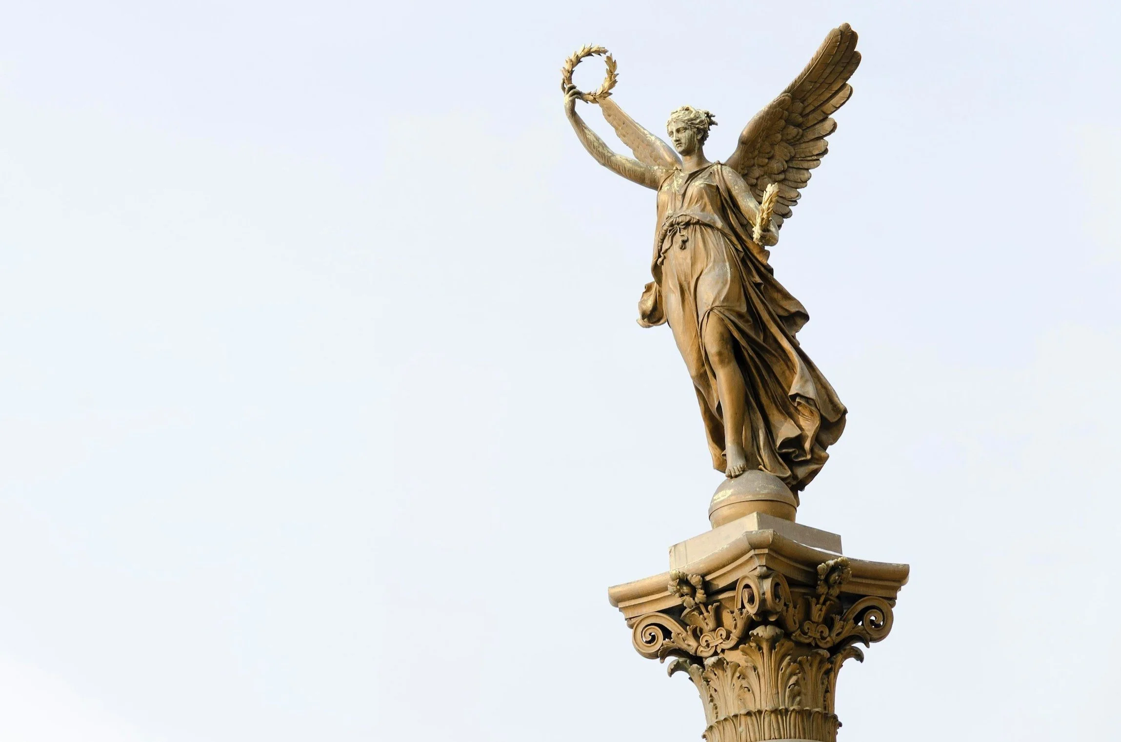 Statue of a winged woman holding a wreath and a scroll, standing on an ornate column against a clear sky.