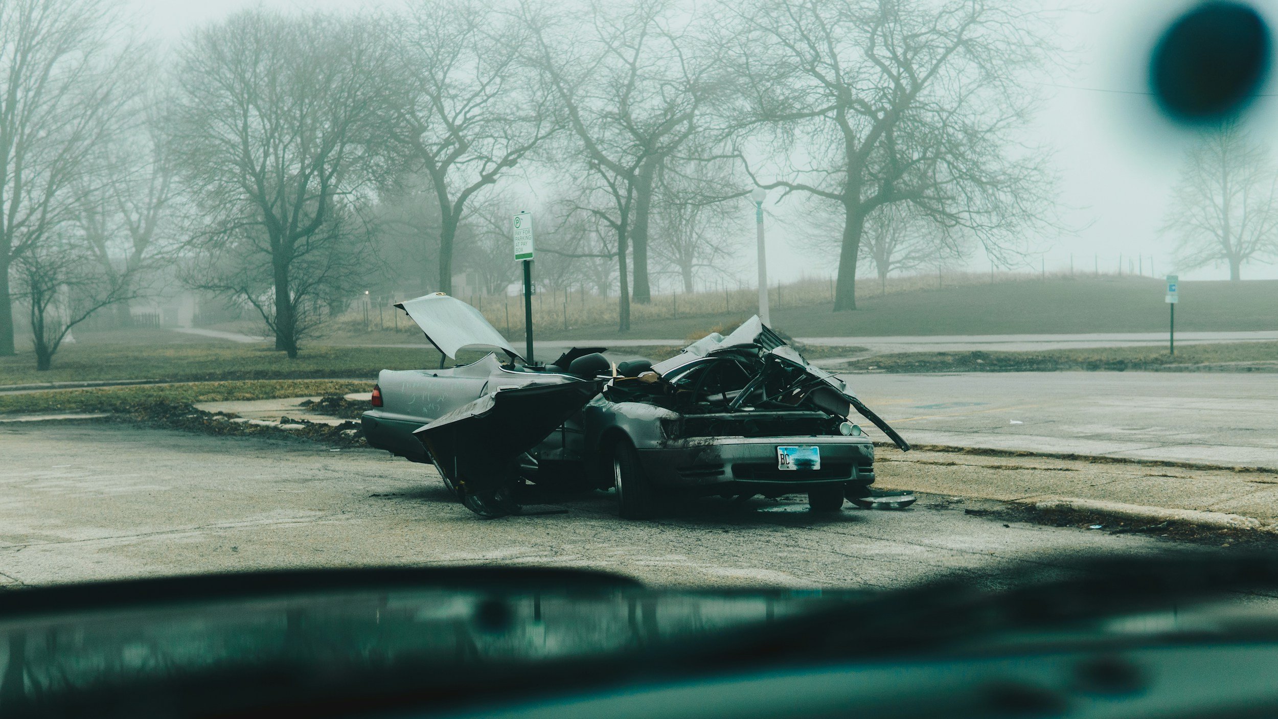 A car crash scene on a foggy day with a severely damaged gray car in the parking lot, airbags deployed, and debris scattered around.