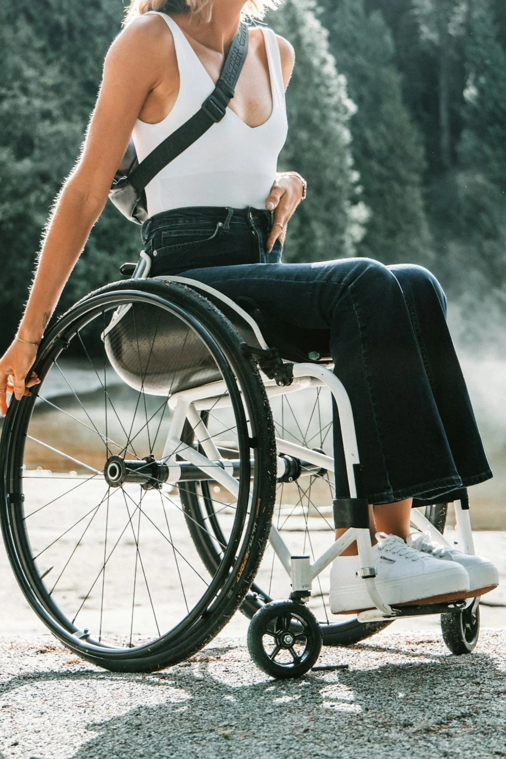 A woman in a white tank top and black jeans sitting in a wheelchair outdoors near a body of water with trees in the background.