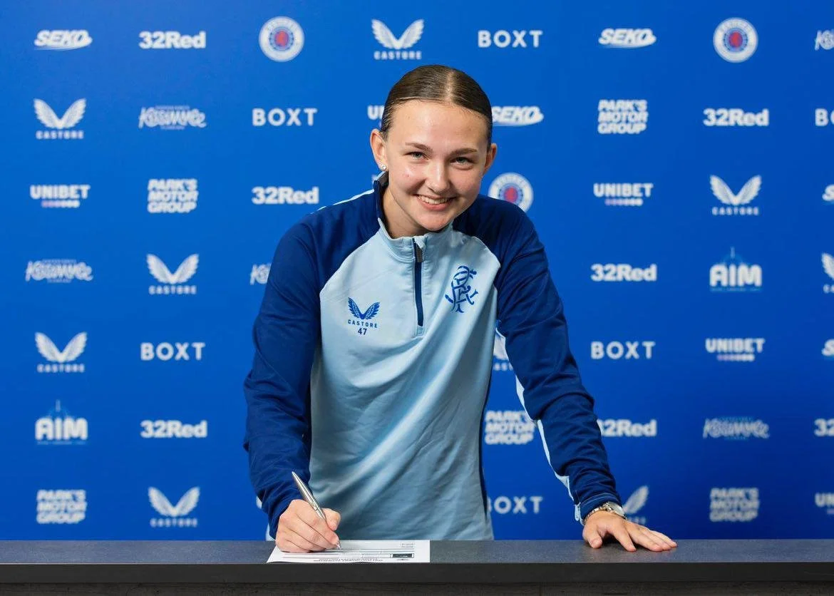 A young woman with slicked-back hair signing a document at a sports event, wearing a blue and light blue sports jacket, in front of a blue backdrop with various sports and company logos.