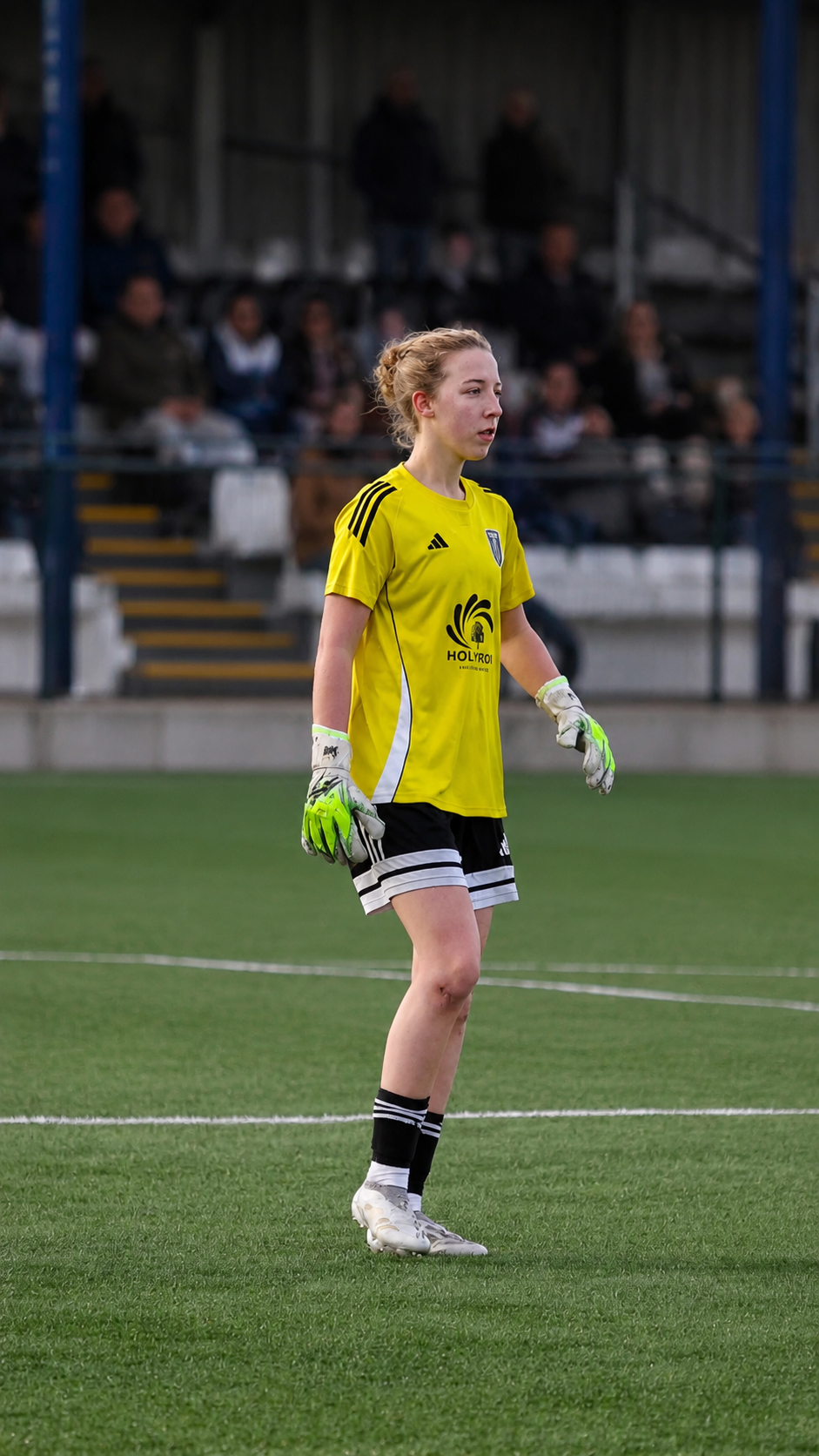 Female soccer goalkeeper standing on the field, wearing a yellow jersey, black shorts, and green goalkeeper gloves, with a crowd of spectators in the background.