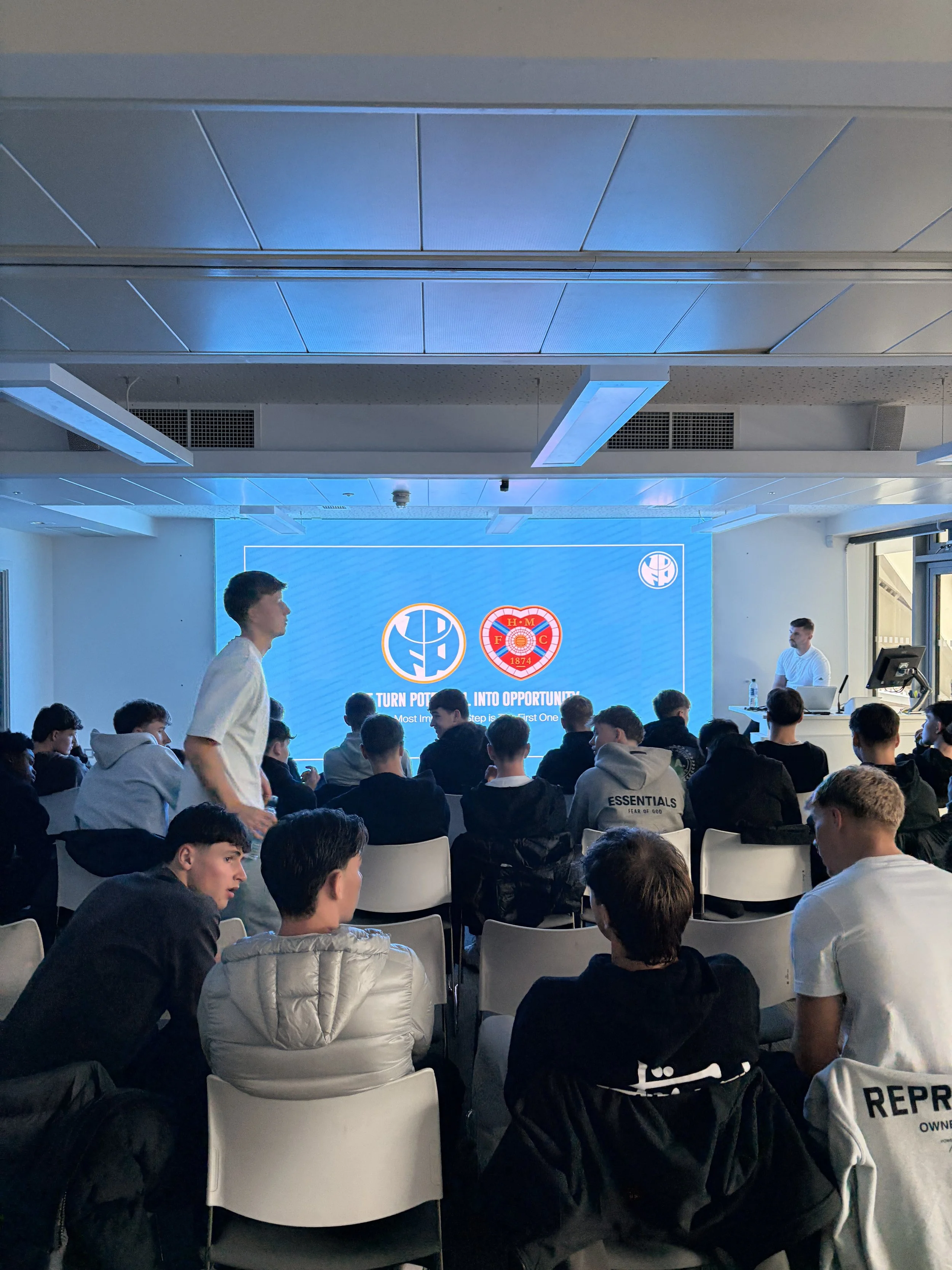 A group of young men sitting in chairs facing a large blue screen with logos and text, during a presentation or event.