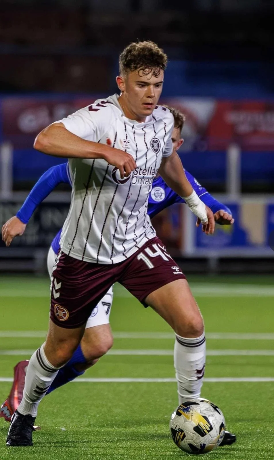 A soccer player in a white and maroon uniform dribbling a soccer ball on the field, with another player in a blue jersey behind him.