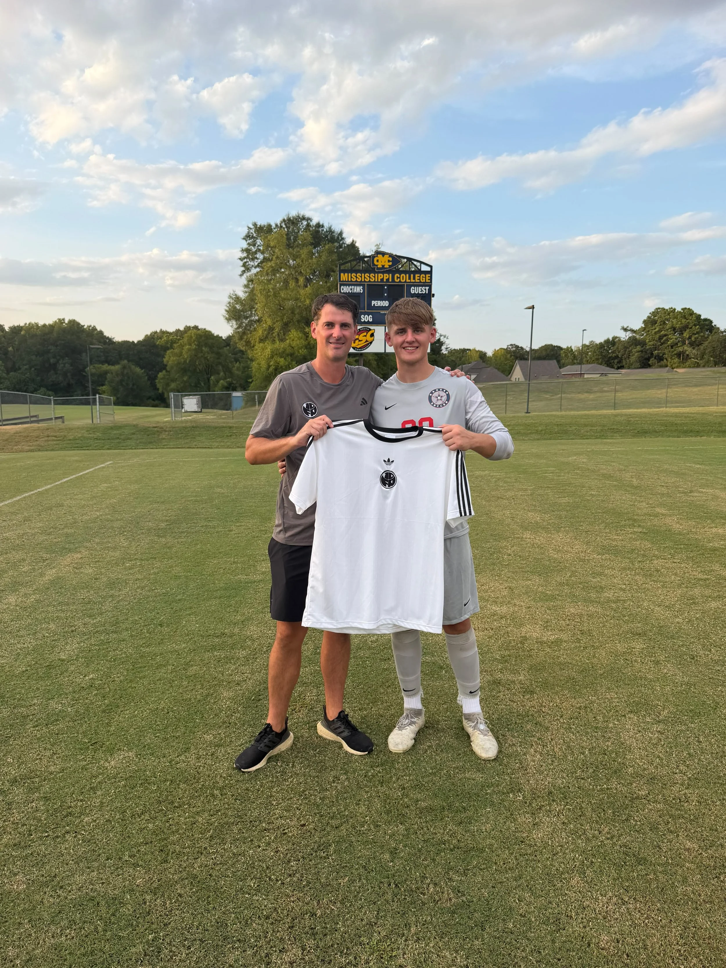 Two men standing on a soccer field, smiling and holding a white soccer jersey. The man on the left is wearing a gray t-shirt and black shorts, and the man on the right is wearing a gray soccer jersey with the number 20, white shorts, and white soccer cleats. They are in front of a scoreboard that reads "Mississippi College" and a blue sky with white clouds.