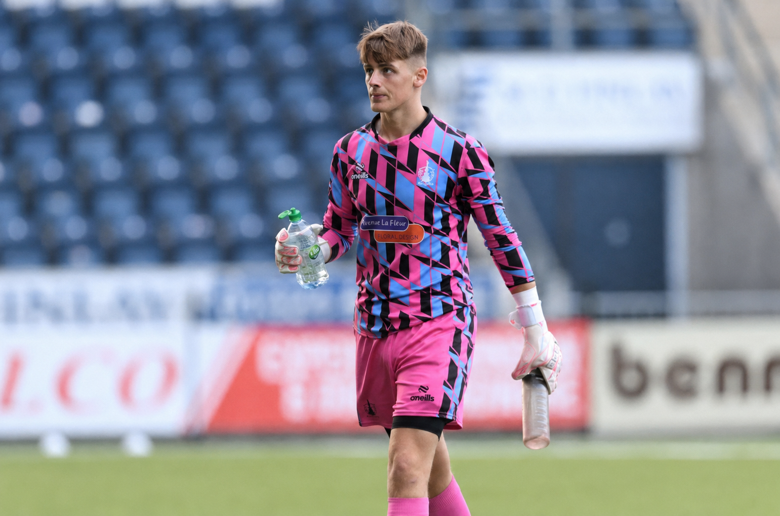 A soccer player during a match wearing pink and blue patterned goalkeeper uniform holding a water bottle and a goalkeeper glove, with empty stadium seats in the background.