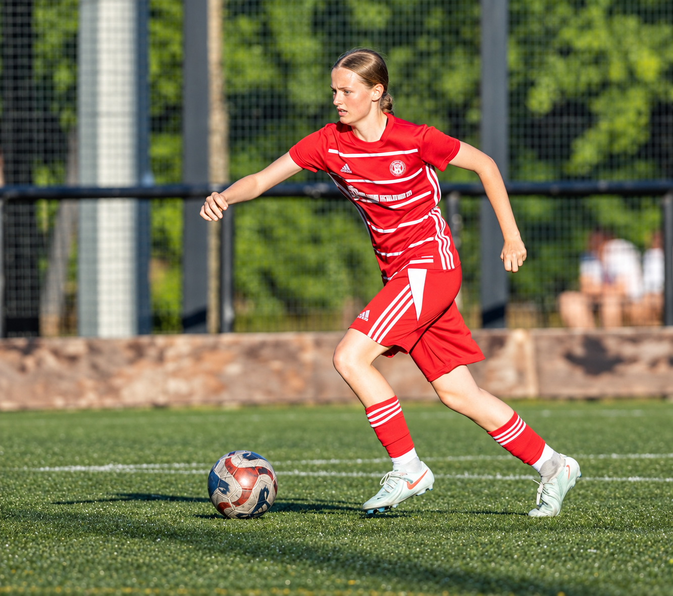 A female soccer player in red uniform dribbling a soccer ball on a green field during daytime.