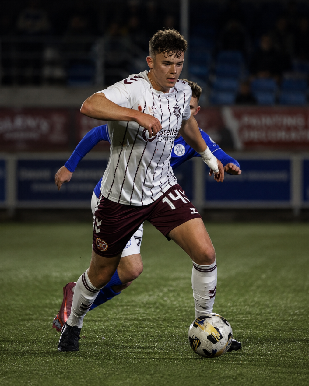 A soccer player in a white and maroon uniform dribbling the ball during a match at night, with an opposing player in blue nearby.